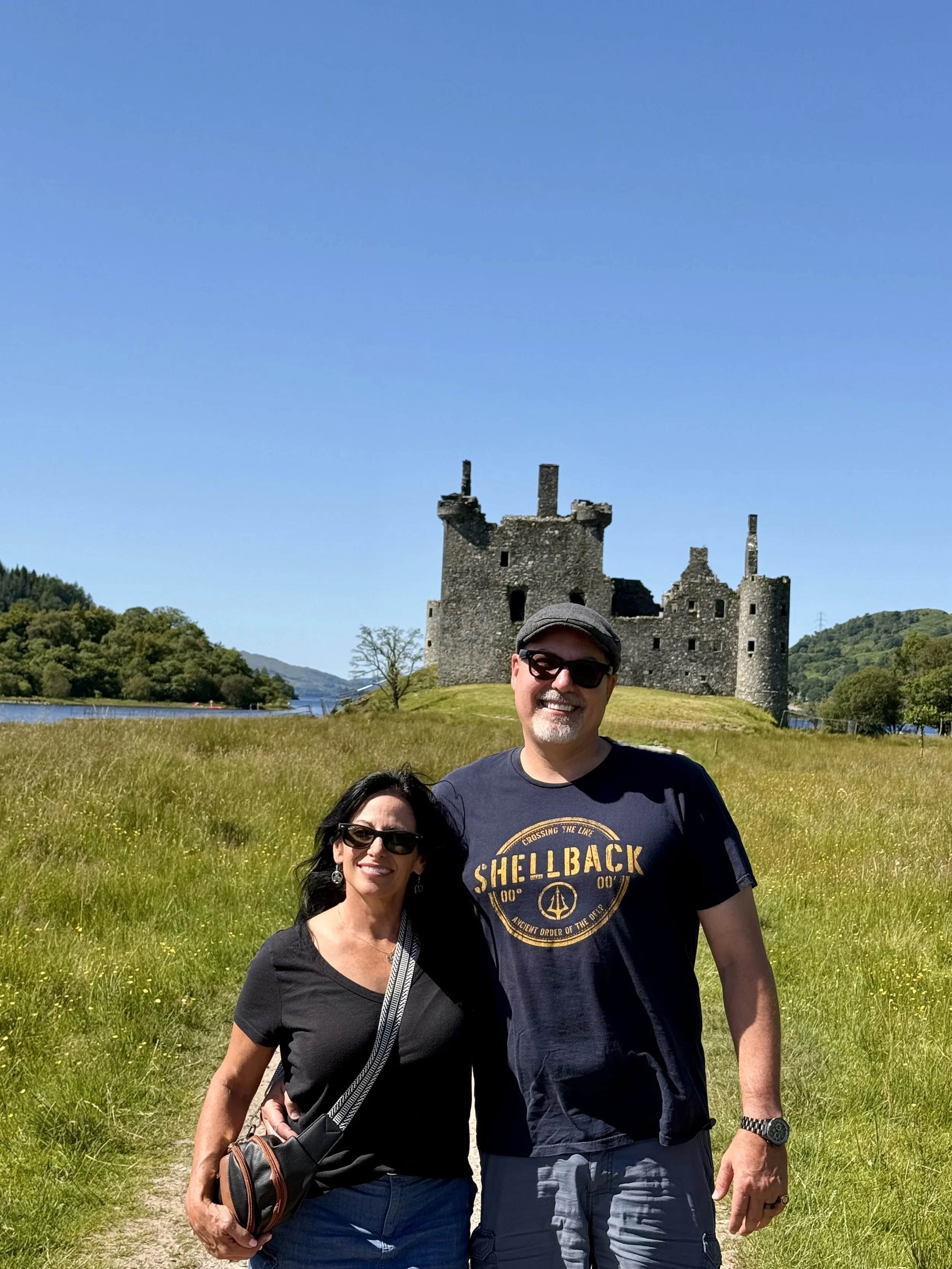 A smiling man and woman standing outdoors on a sunny day, with a castle ruins in the background and lush green rolling hills around. The man is wearing a black T-shirt with yellow text, sunglasses, and a watch. The woman is wearing a black shirt and sunglasses, with a shoulder bag.