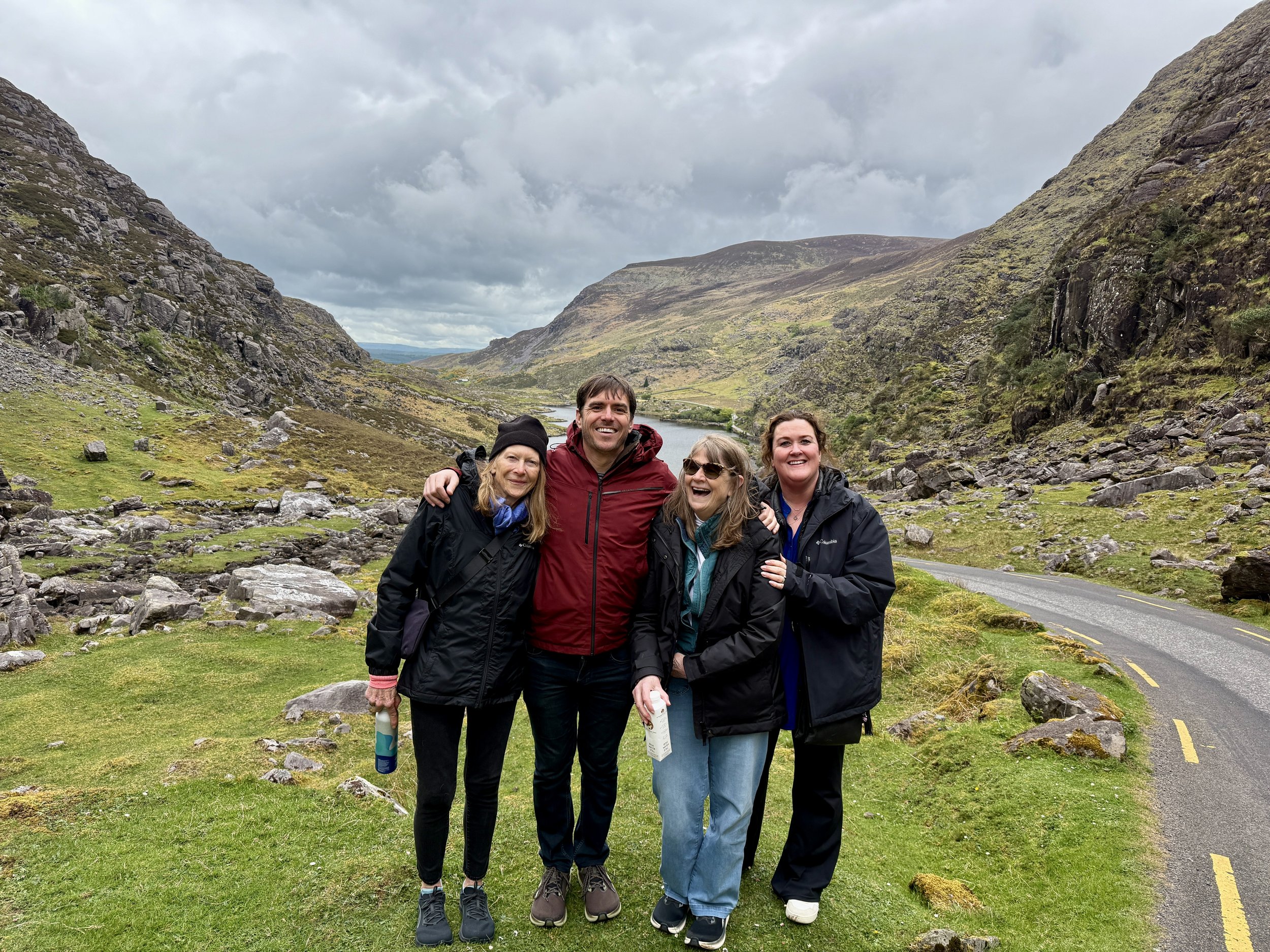 Four people standing together outdoors in a mountainous landscape, smiling with rocky slopes and cloudy sky in the background.
