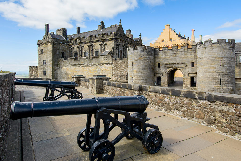 Two cannons on a stone walkway facing a historic stone castle with battlements and towers, under a partly cloudy sky.