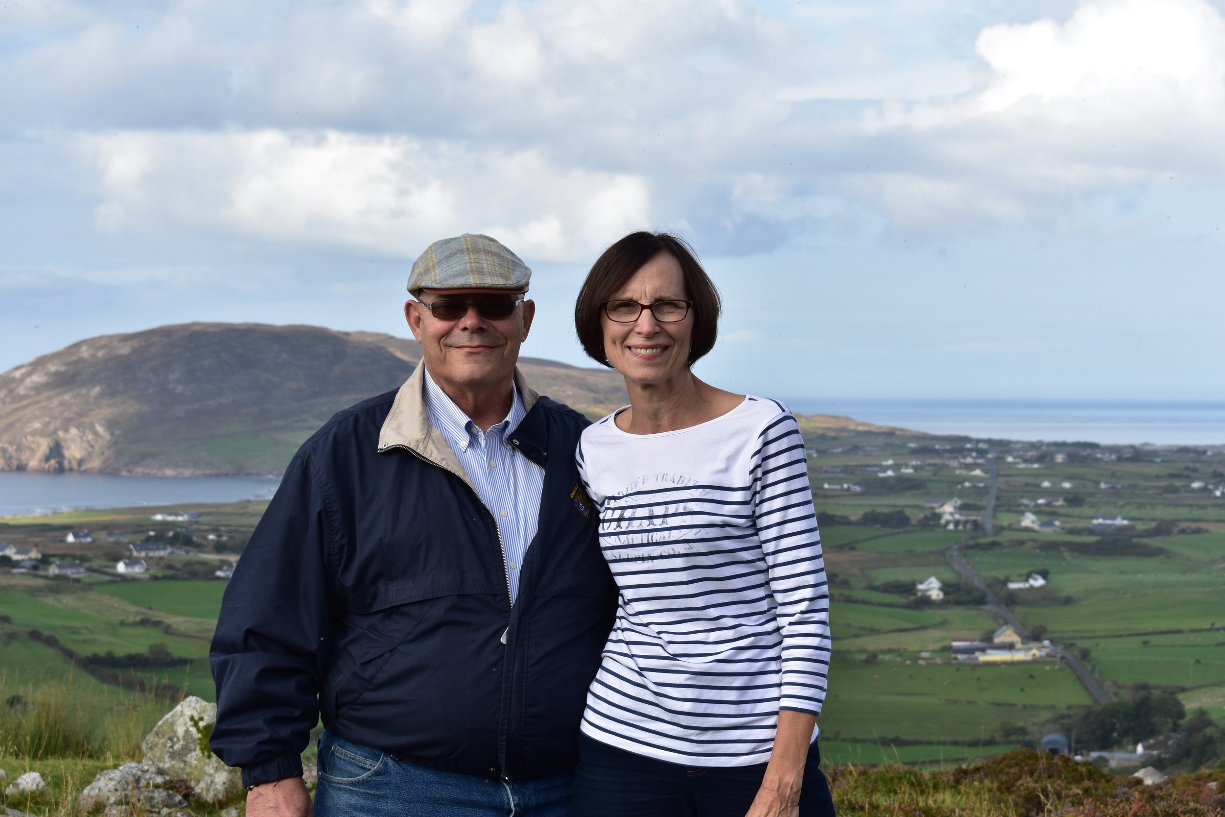 A middle-aged man and woman standing outdoors on a cloudy day with a scenic backdrop of hills, water, and farmland. The man is wearing a gray cap, sunglasses, a light blue shirt, and a black jacket. The woman has short dark hair, wears glasses, and a striped shirt.