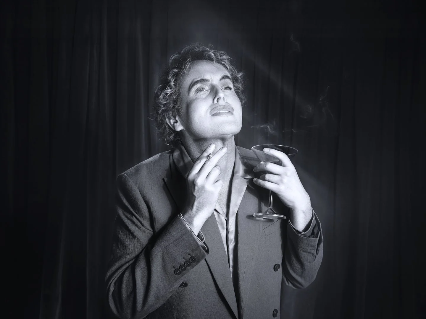 A black and white photo of a woman in a suit, holding a cocktail glass, looking upward with a relaxed, confident expression, in front of a dark curtain background.