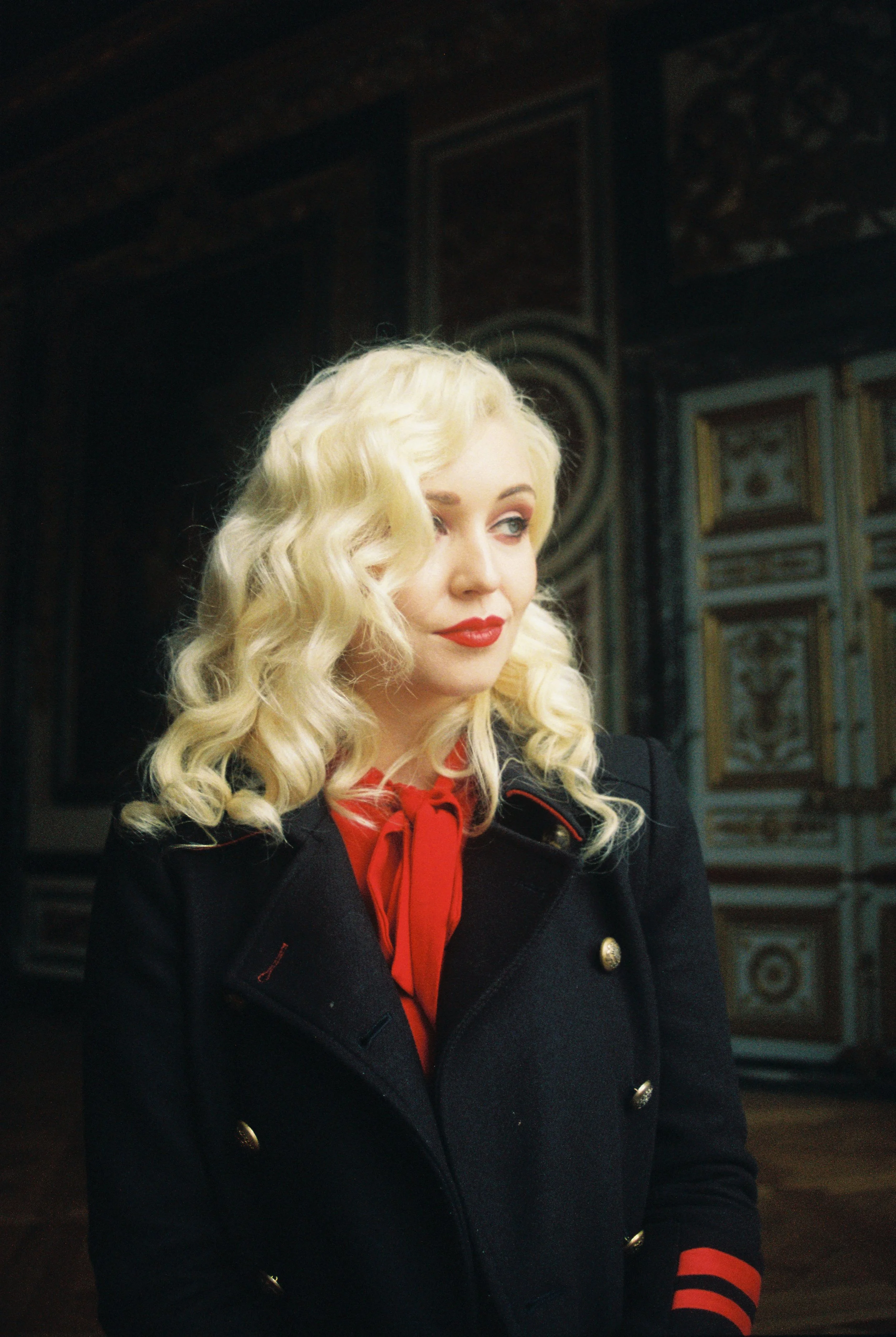 A woman with platinum blonde, wavy hair styled in vintage curls, wearing a black coat with gold buttons, a red blouse with a bow tie, and red and black striped cuffs. She is standing indoors with ornate, framed artwork in the background.