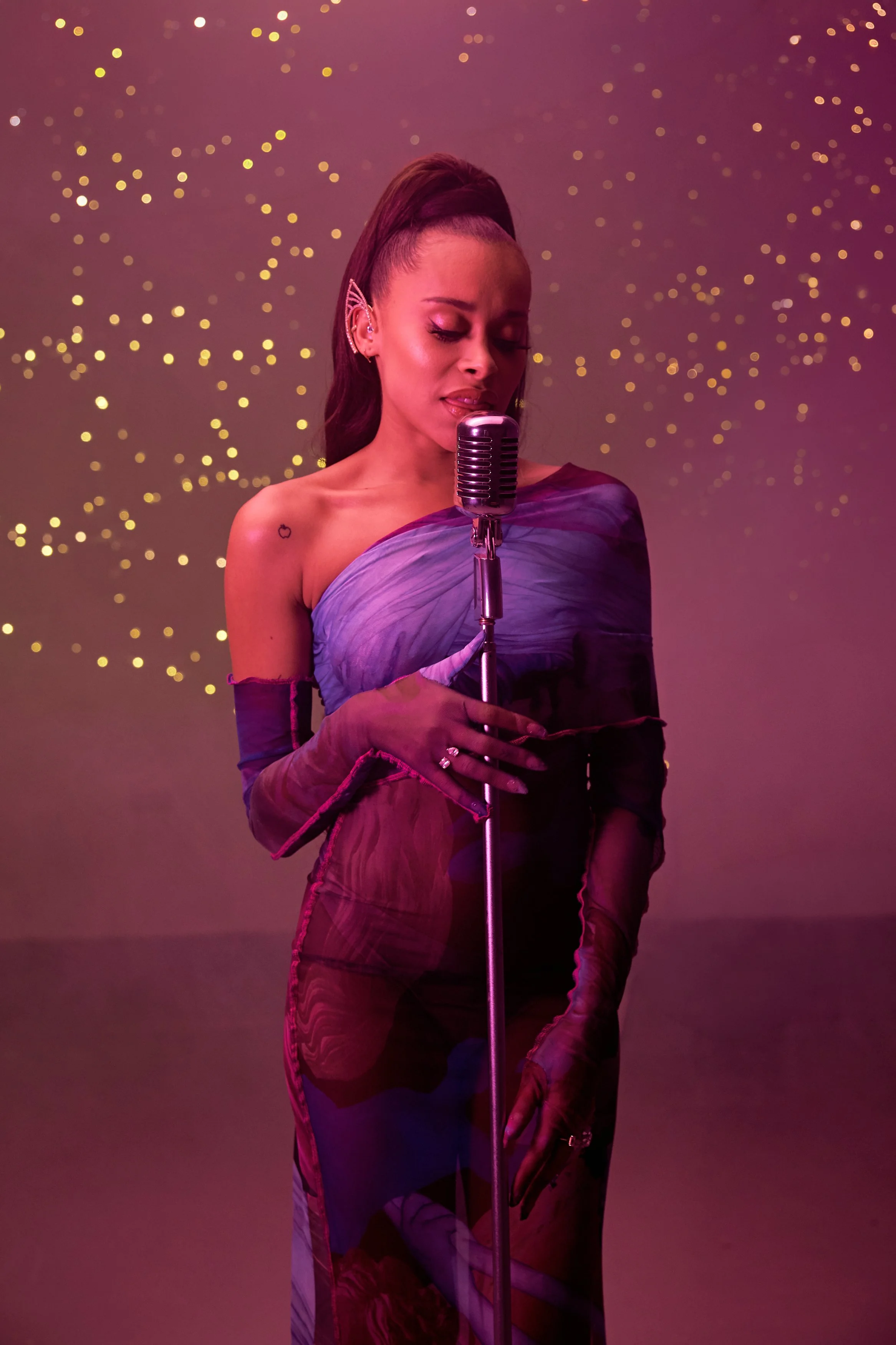 A woman singing into a vintage microphone against a backdrop of soft pink and purple lighting with small glowing dots.