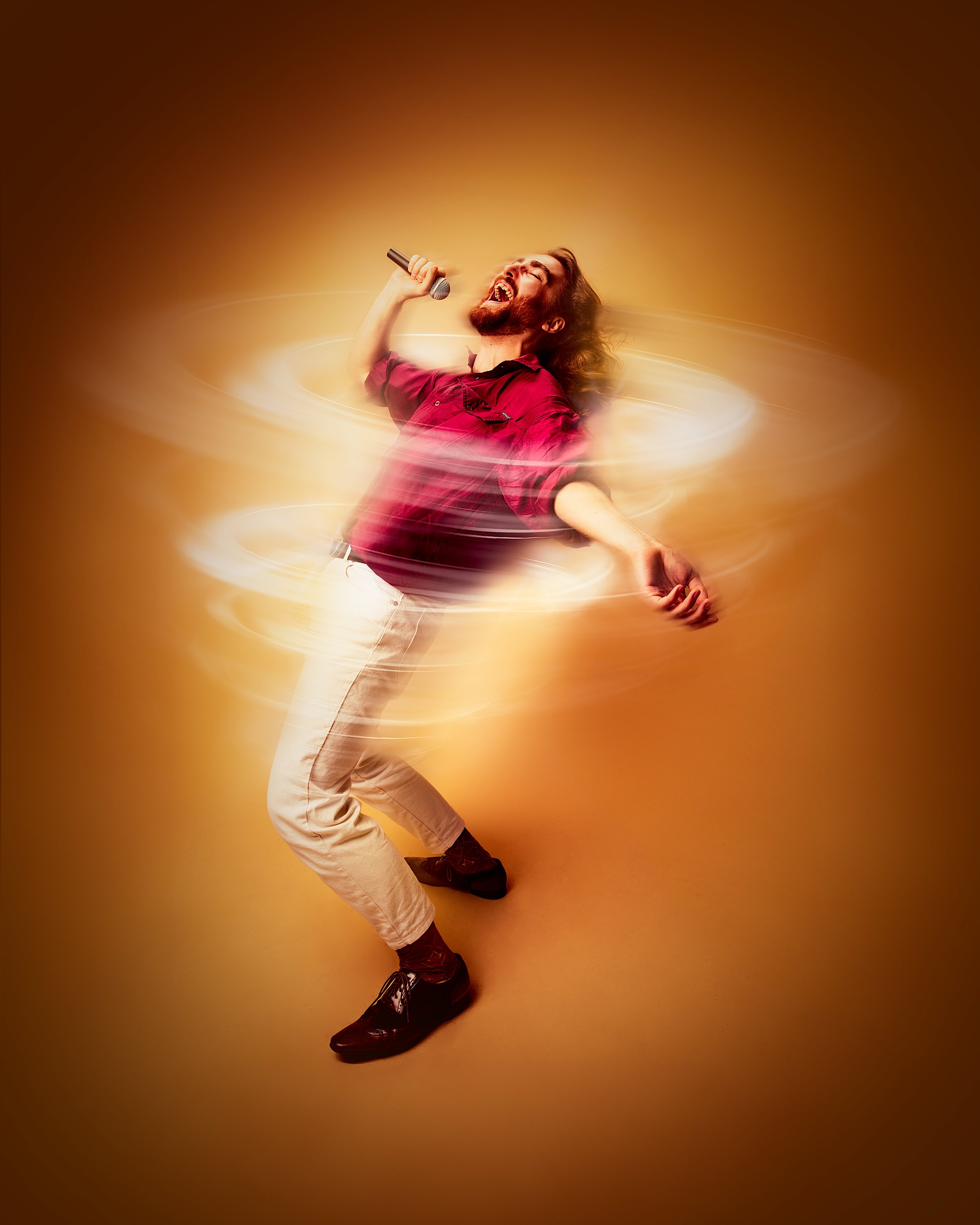 A man with long hair and a beard singing passionately into a microphone, wearing a red shirt and light-colored pants, surrounded by swirling light effects against a warm, gradient background.