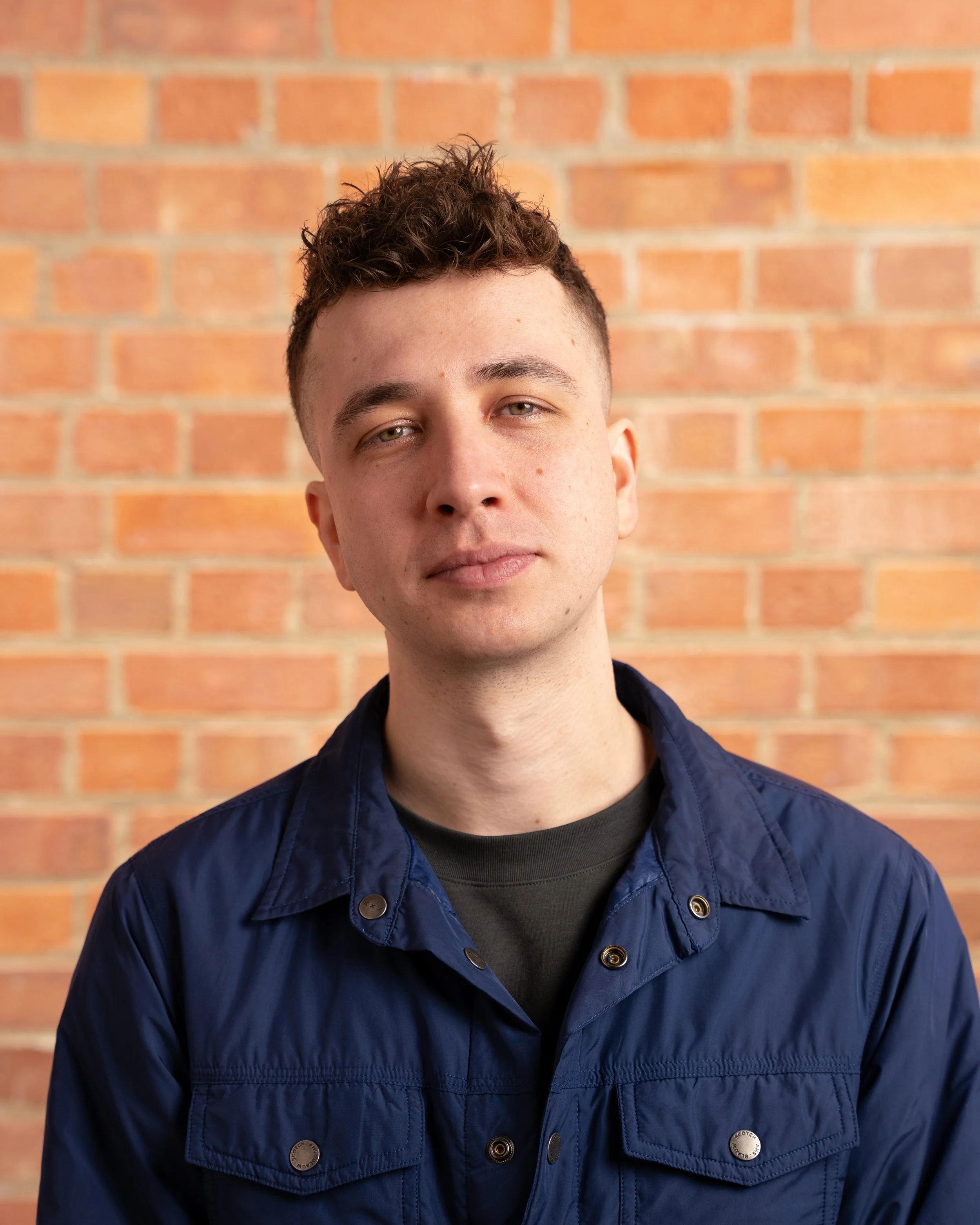 A young man with short curly brown hair and fair skin standing in front of a brick wall. He is wearing a dark shirt underneath a navy blue jacket with shiny buttons.