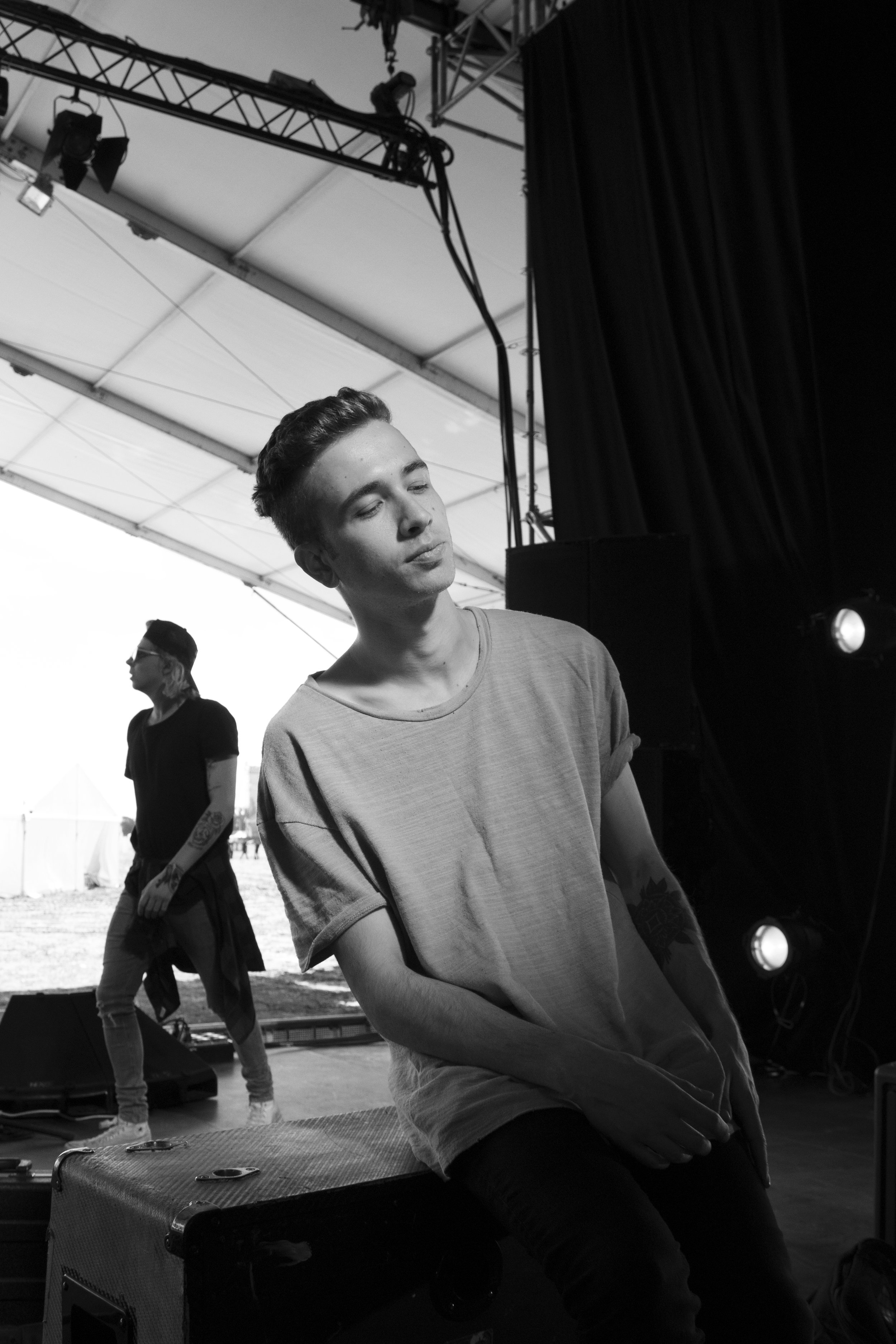 A black and white photo of a young man with short hair, closed eyes, and a relaxed expression, sitting on a ledge. Behind him are other people and stage equipment under a tent or canopy.