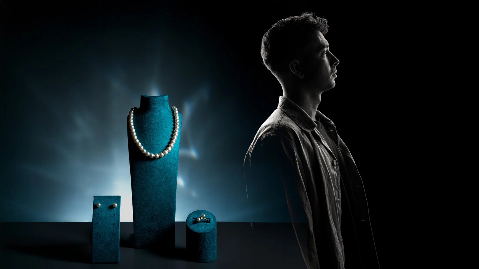 A man in profile looking to the right, with a dark background and a display of jewelry including a pearl necklace, earrings, and rings on blue velvet stands.