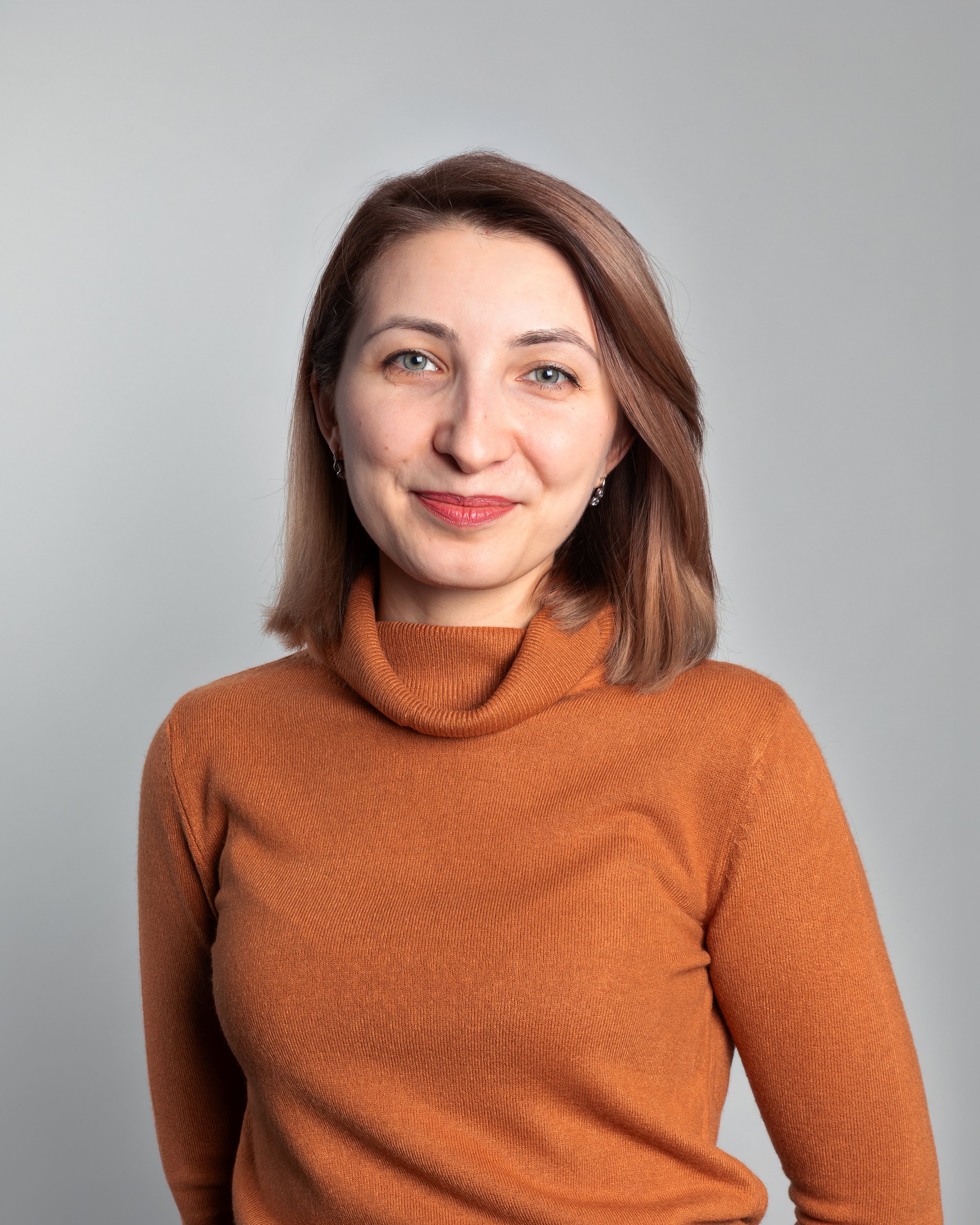 A young woman with light brown hair and blue eyes, wearing an orange turtleneck sweater, smiling against a plain light gray background.