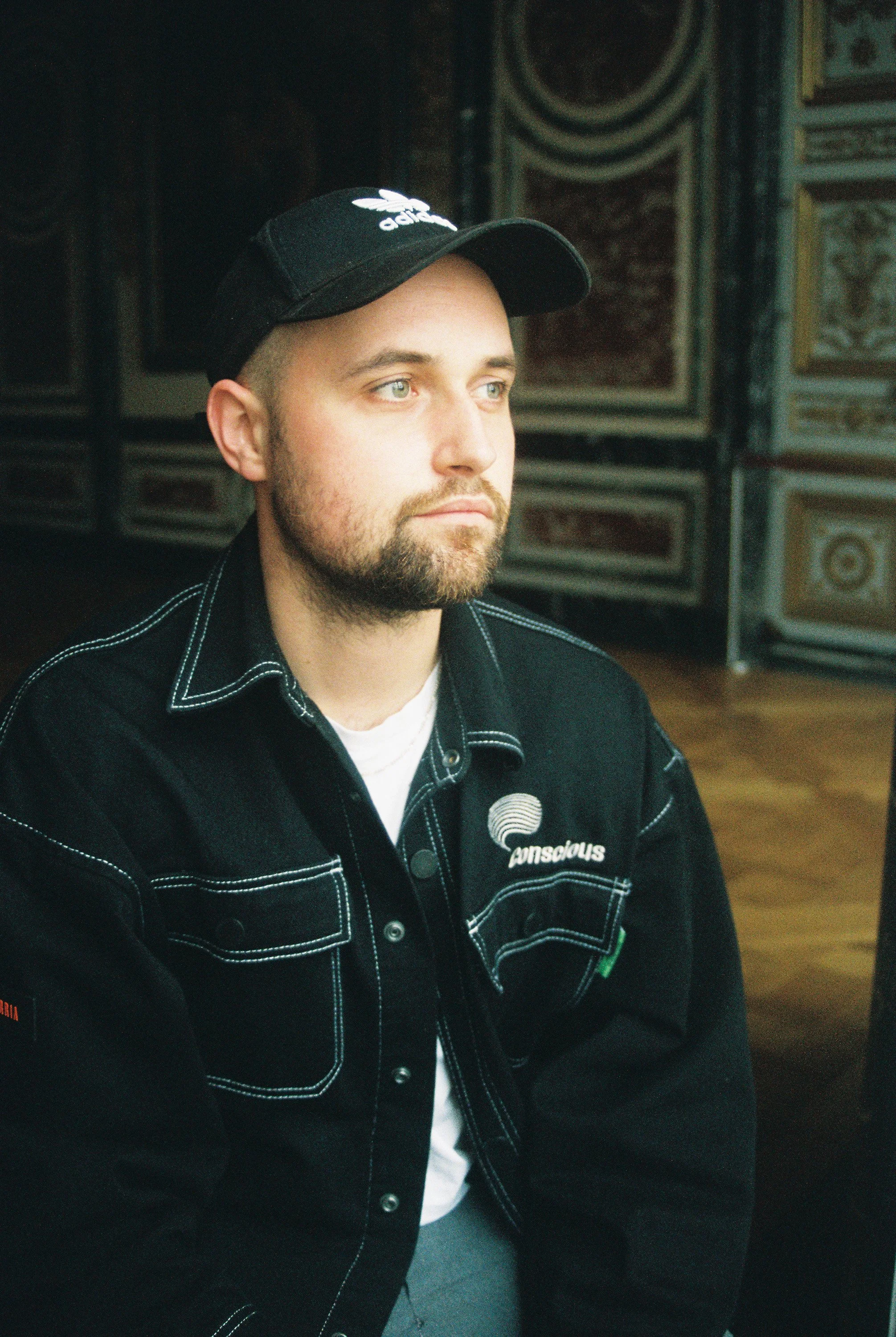 A young man with light skin, blond hair, and a beard, wearing a black Adidas cap and a black denim jacket with the word 'Conscious' embroidered on it, seated indoors with a decorative wall in the background.