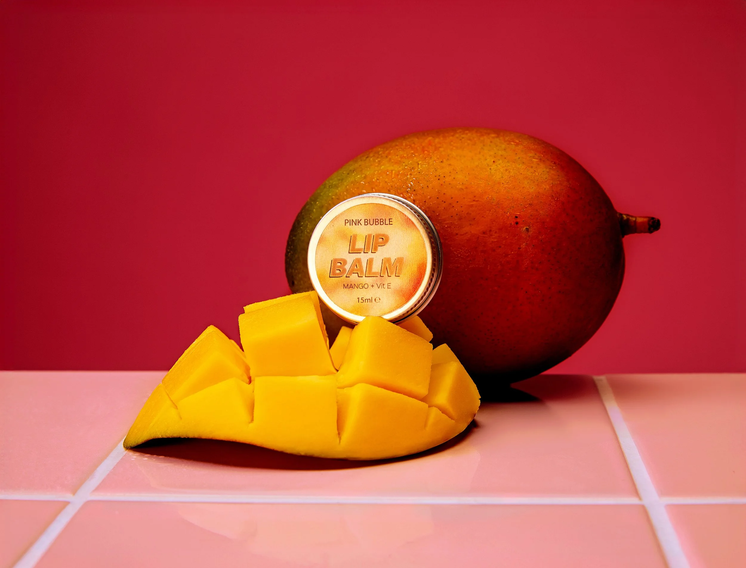 A cut mango and a jar of pink bubble lip balm with mango and vitamin E on a pink background.
