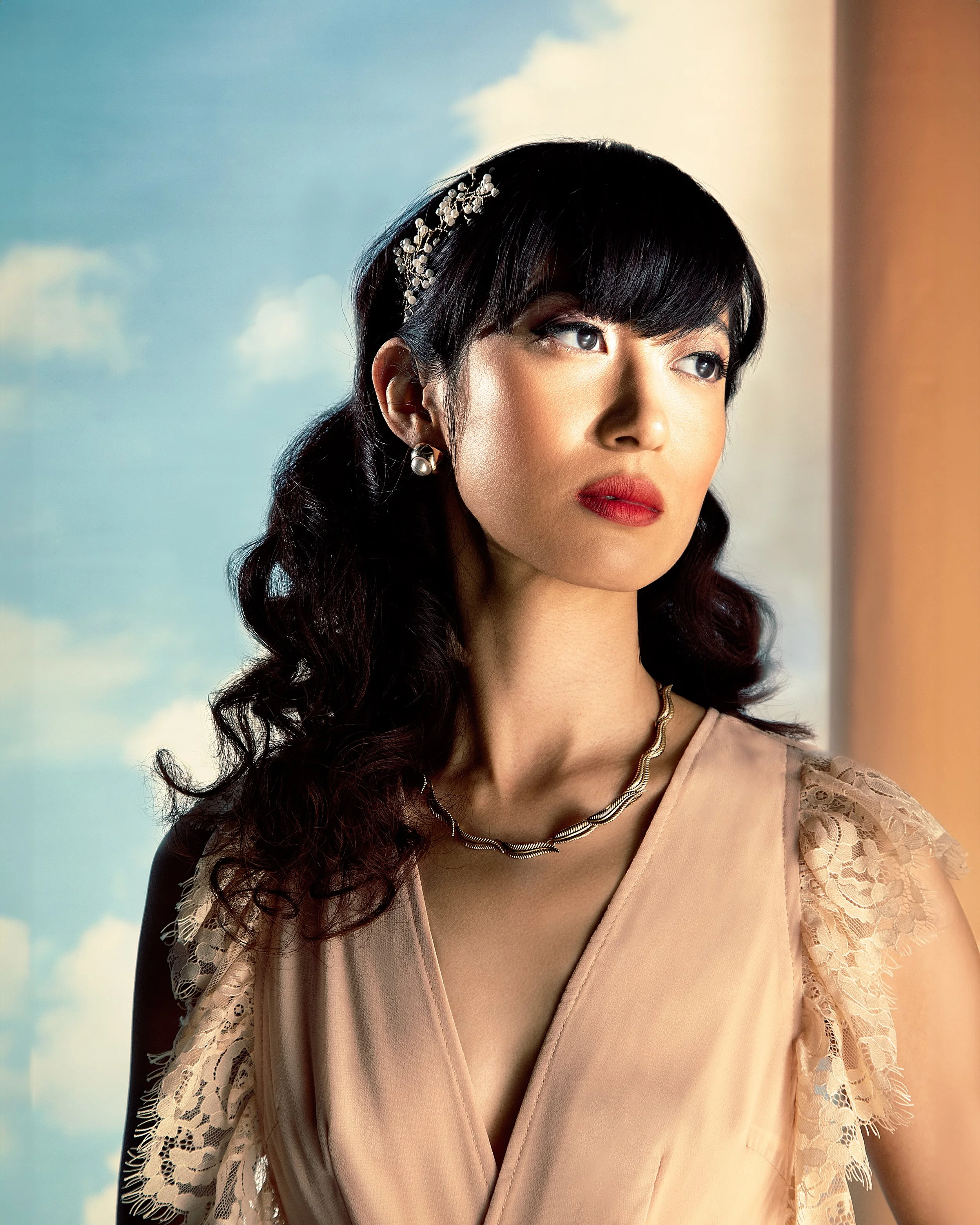 Close-up of a woman with dark hair, wearing earrings with pearls and ornate hair accessory, dressed in a peach-colored dress with lace details on the sleeves, looking thoughtfully into the distance against a background of a blue sky with clouds.
