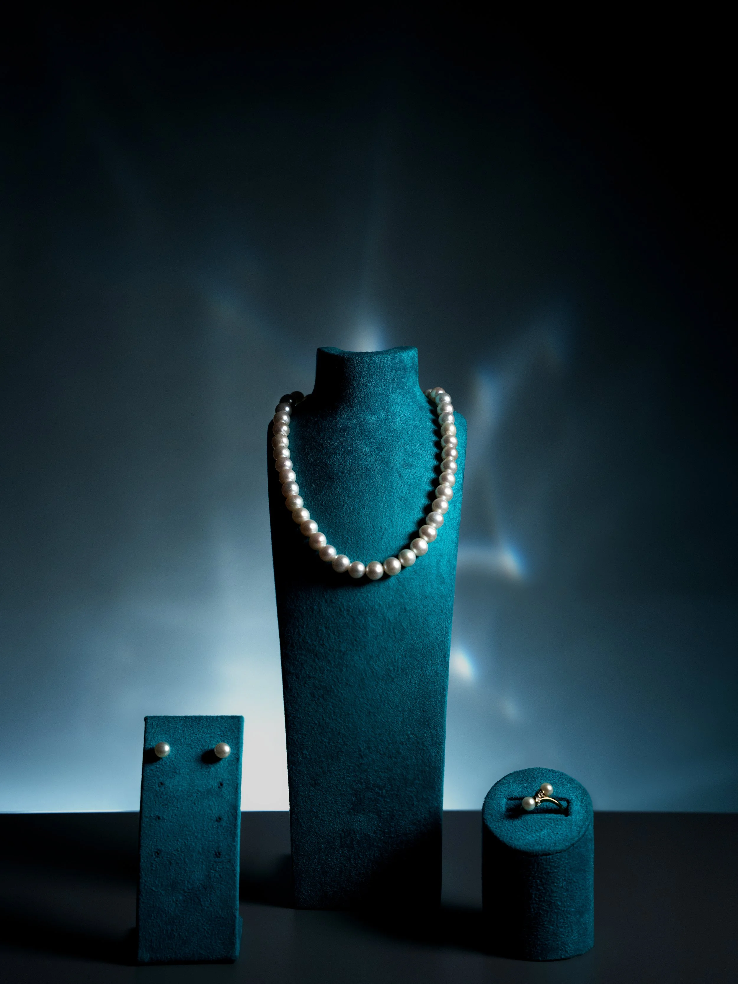 Jewelry display with a string of pearls on a blue velvet stand, matching pearl earrings, and a pearl ring, all on blue velvet stands against a dark background.