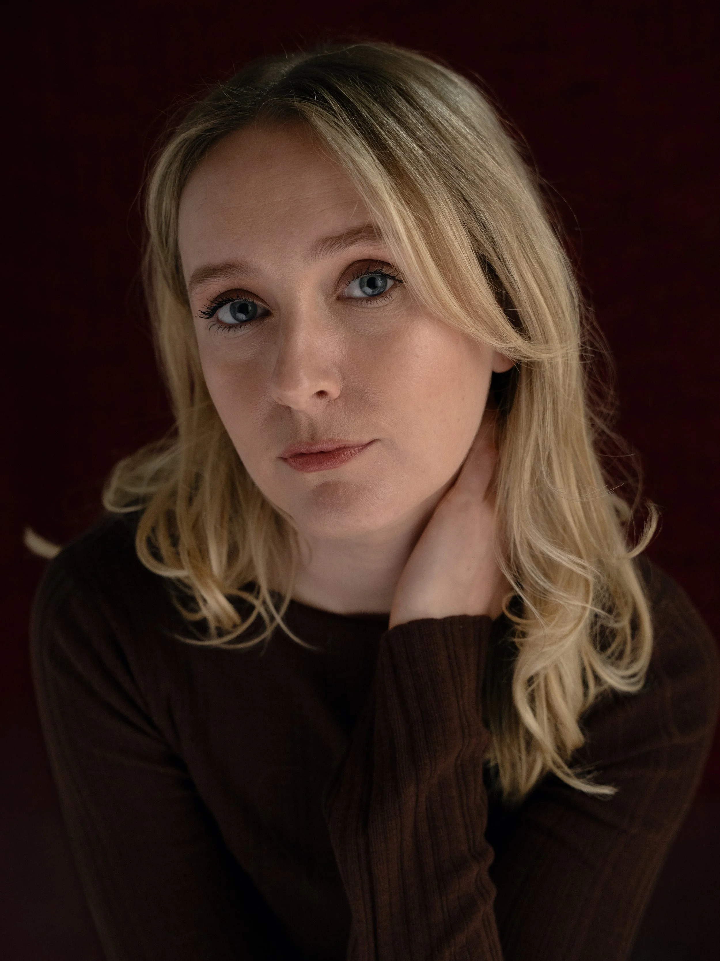 A young woman with blonde hair and blue eyes looking at the camera, wearing a dark sweater, in a dimly lit setting.