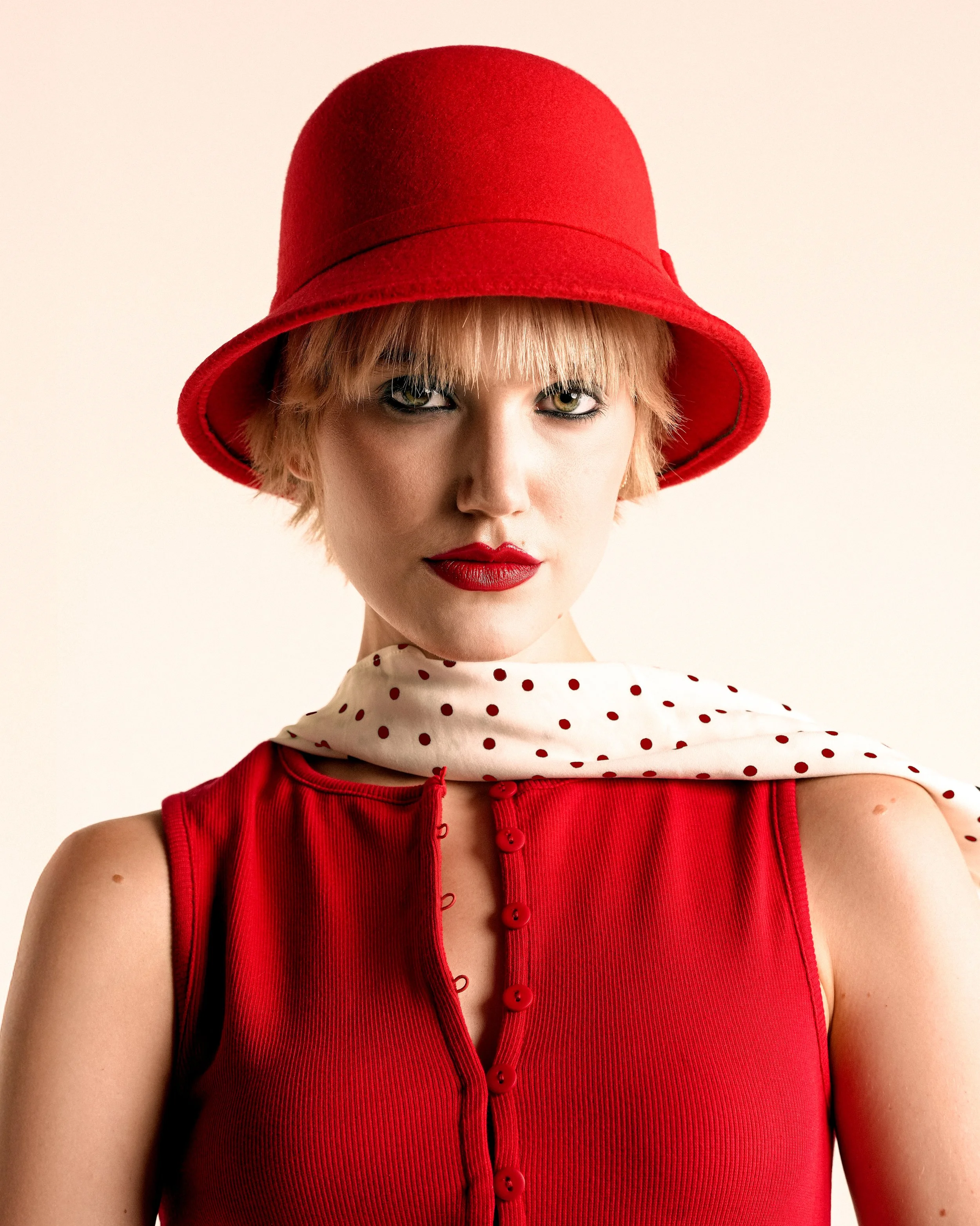 A woman wearing a red hat, red sleeveless top with buttons, and a white scarf with red polka dots.