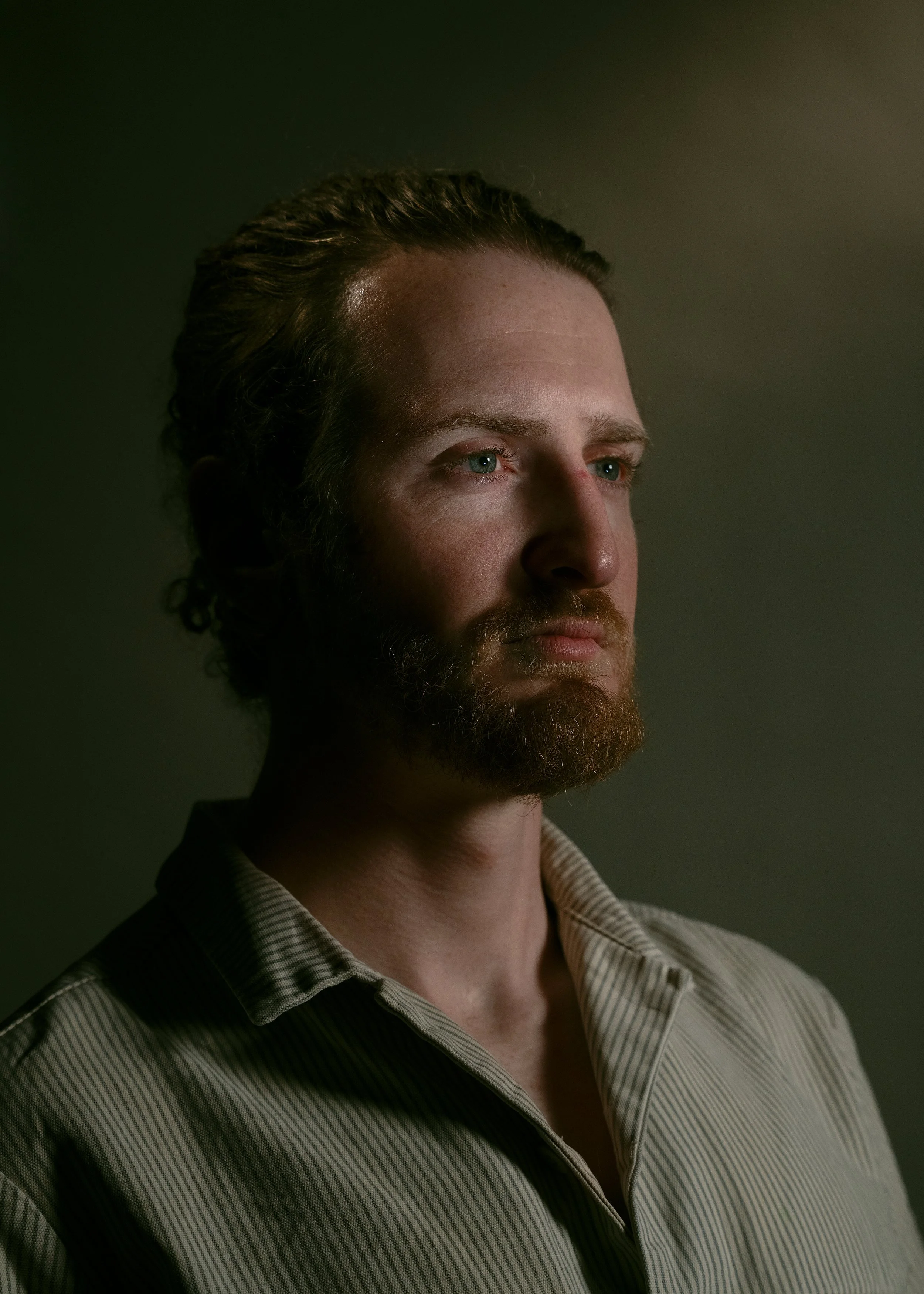 A portrait of a young man with a beard, light skin, blue eyes, and brown hair, wearing a striped collared shirt, looking slightly to the right against a dark background.
