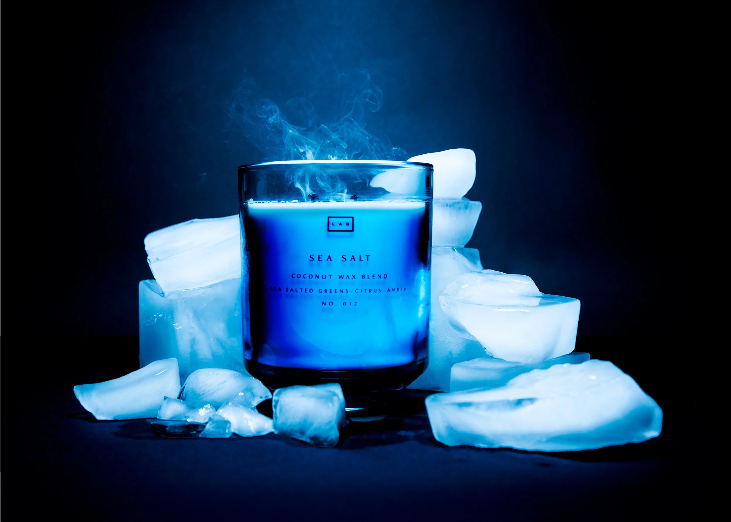 A lit blue candle labeled 'Sea Salt' surrounded by ice cubes and ice blocks against a dark background.