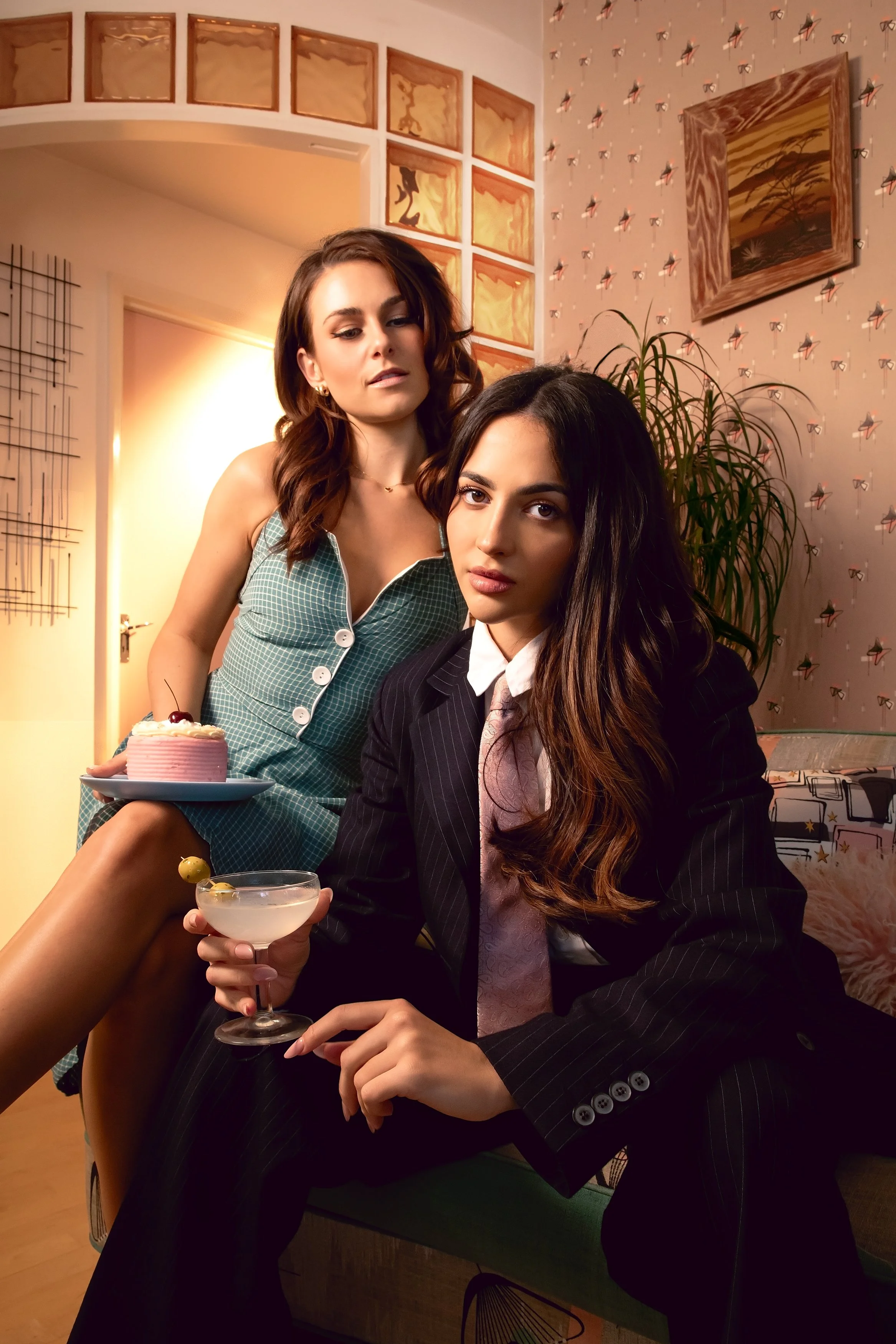 Two women, one in a striped suit with long wavy hair holding a cocktail glass, and the other in a checkered dress with shoulder-length hair, are in a room with patterned wallpaper, artwork, and indoor plants. The woman in suit is seated, the other standing behind her, and there is a pink cake on a plate and a drink in the foreground.