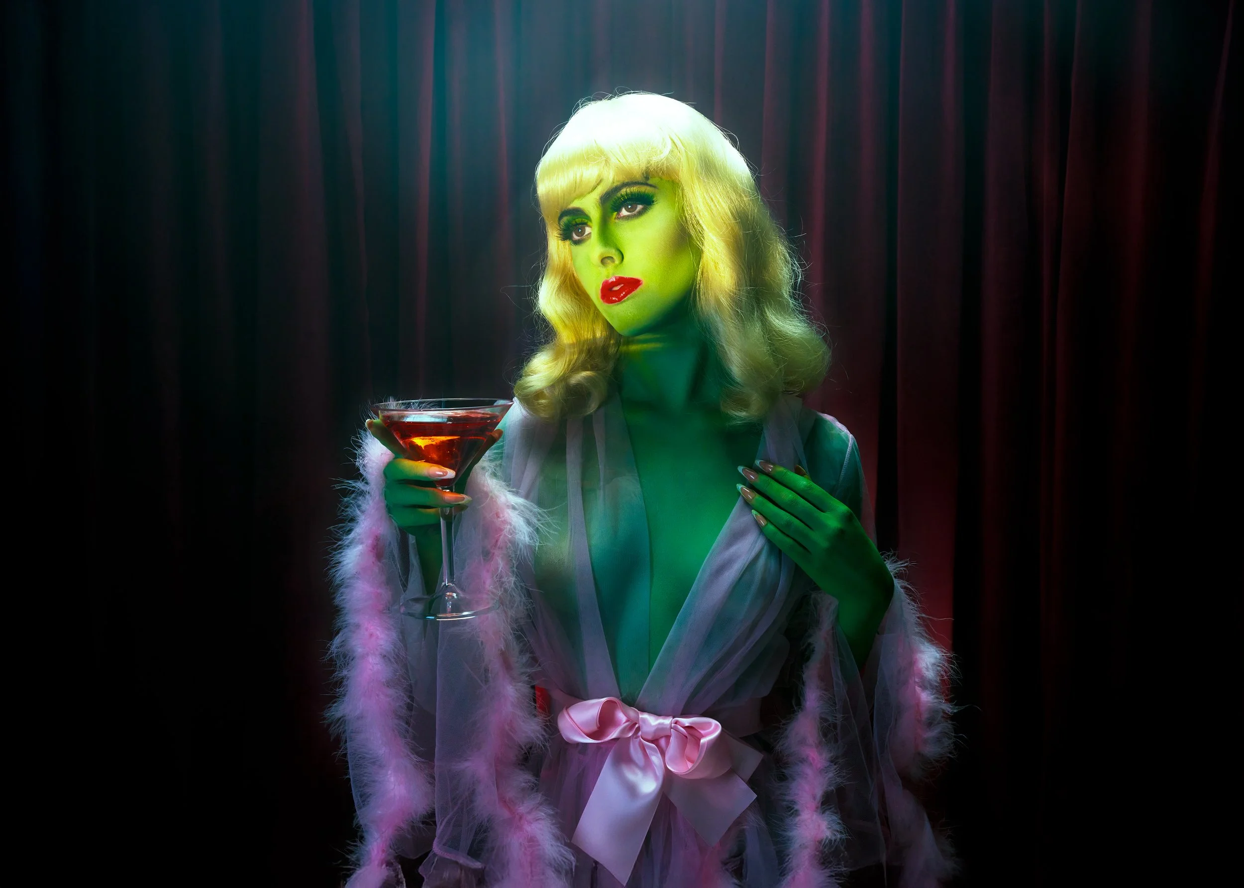 A woman with blonde hair, wearing makeup, holding a martini glass with a red drink, standing in front of dark curtains, dressed in a sheer robe with a pink satin bow and pink feathered trim.