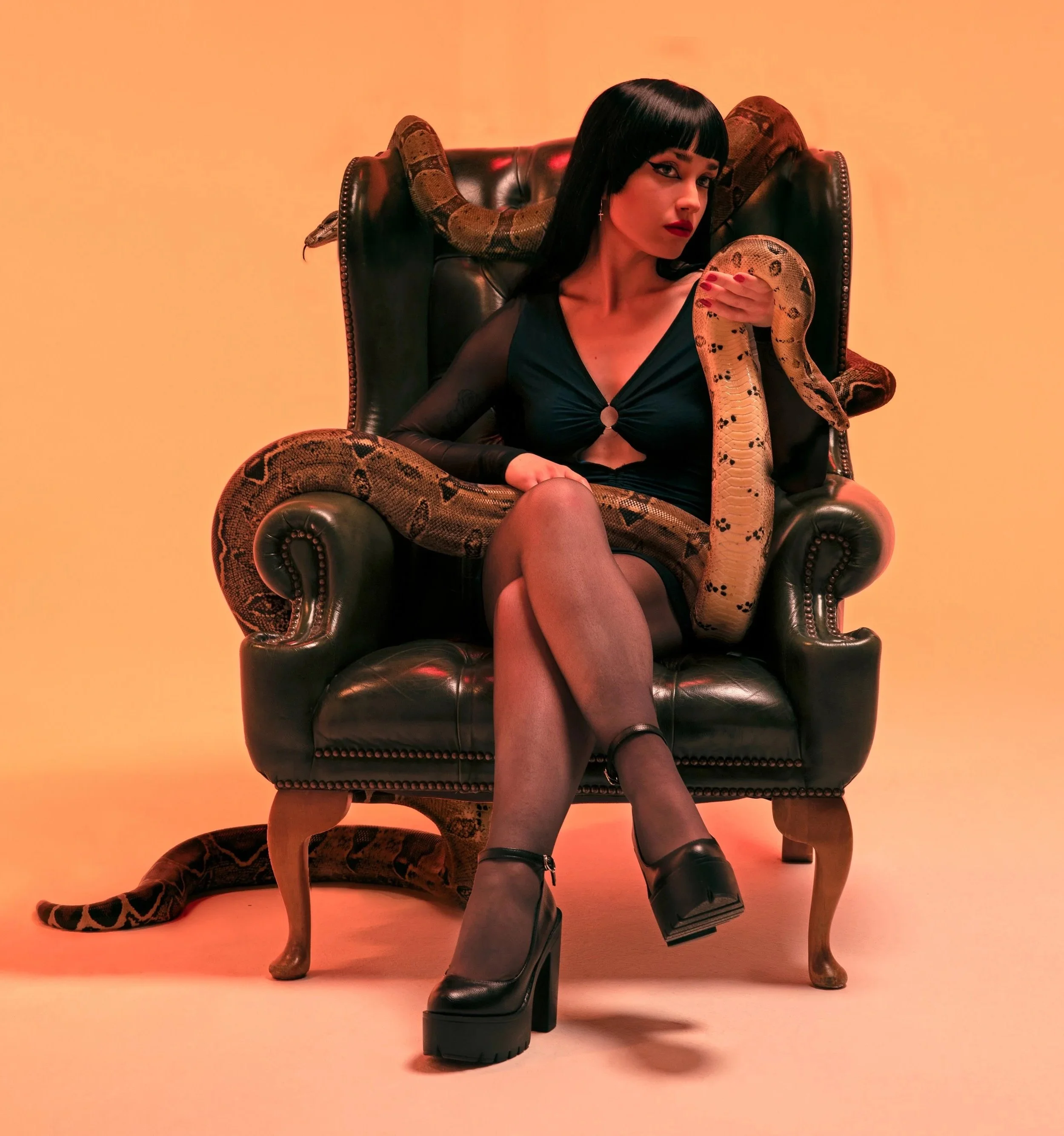 A woman with black hair and red lipstick sitting on a black leather chair with wooden legs, holding a snake on her shoulder and arm, against a peach-colored background.