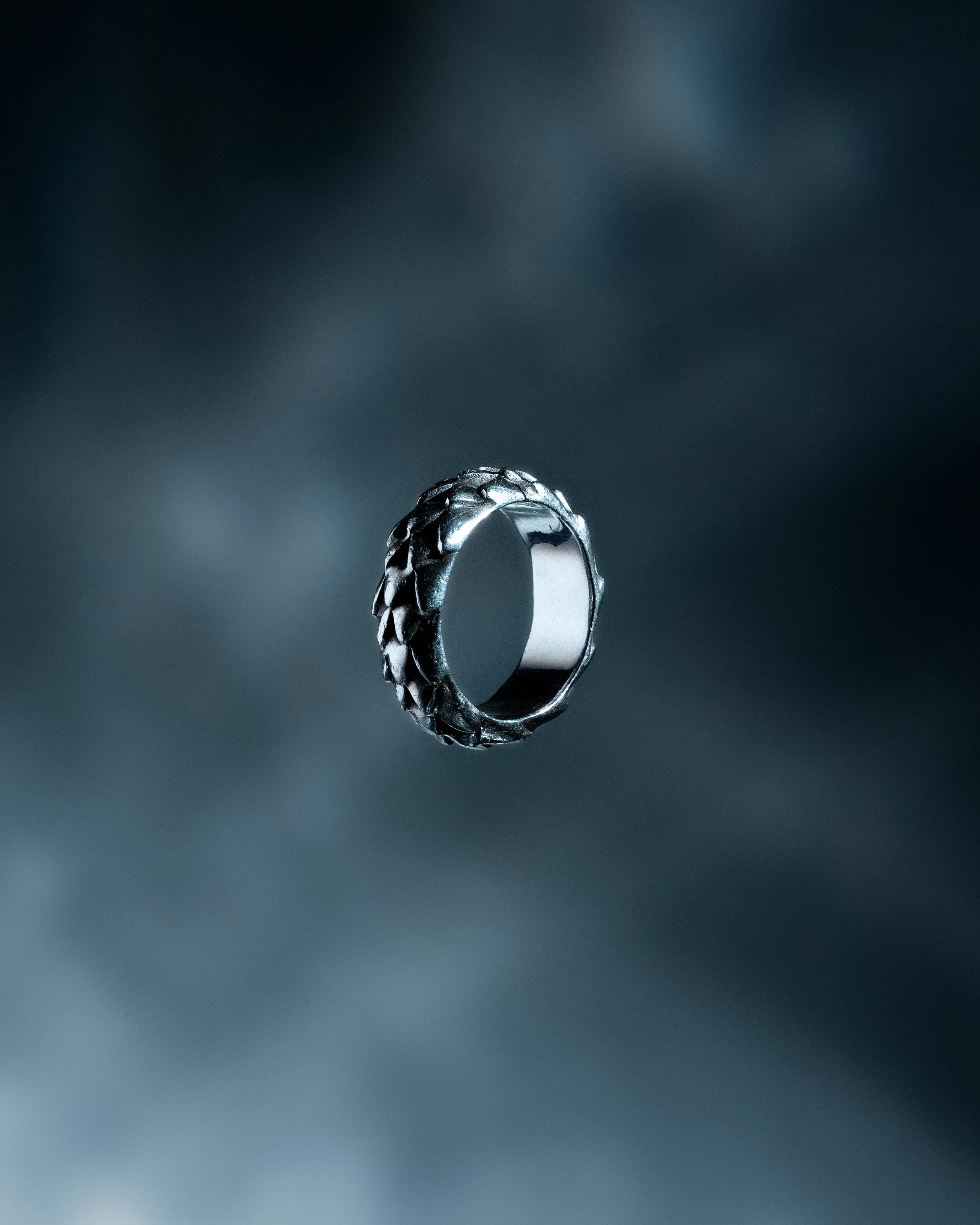 Silver ring with a textured band, floating against a dark, blurred background.