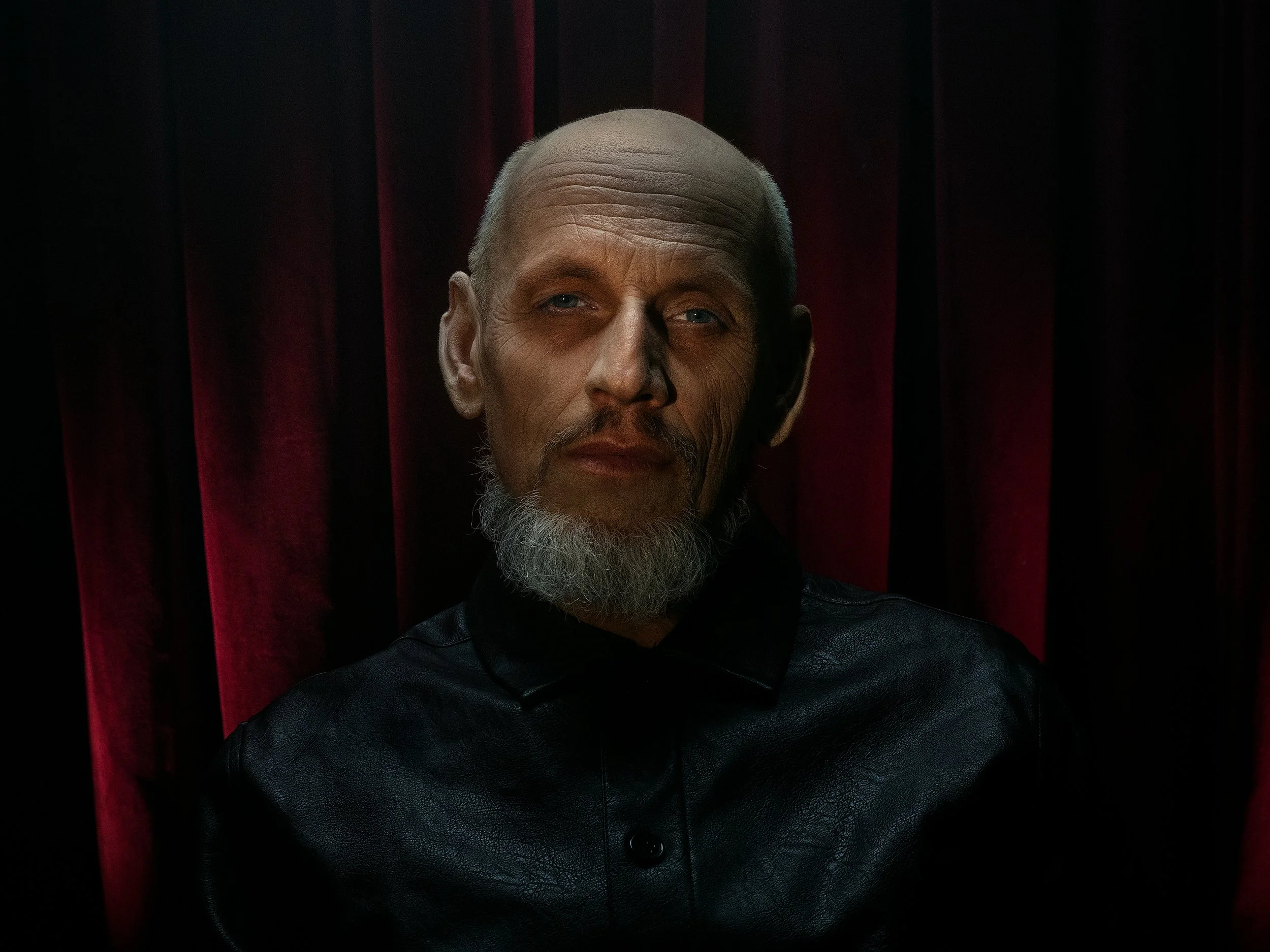 A portrait of a middle-aged man with a shaven head, gray goatee, and piercing eyes, wearing a black leather jacket, set against a dark background with red curtain-like elements.