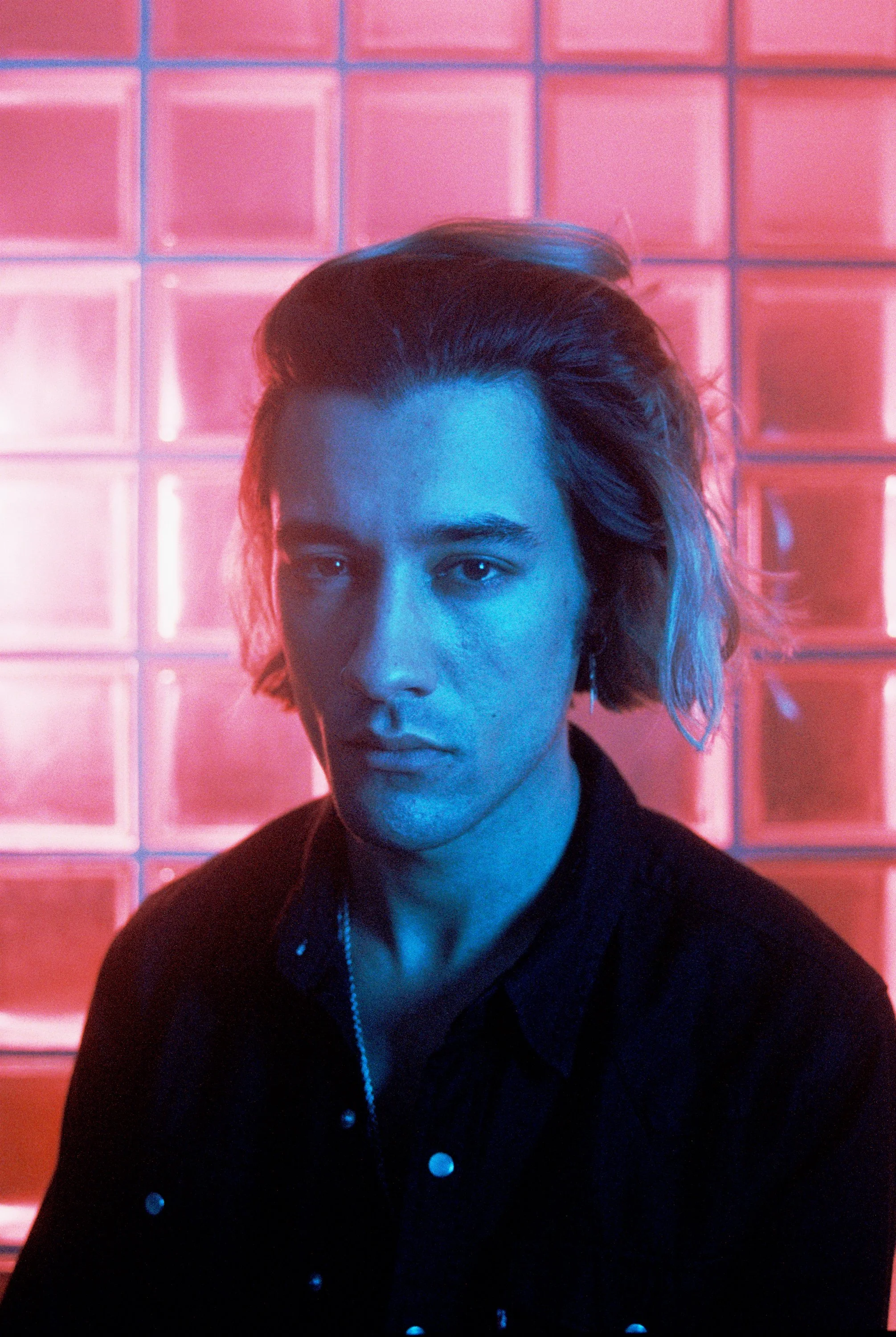 A person with short hair and earrings, wearing a black jacket with silver buttons, standing in front of a wall made of illuminated glass blocks, with pink and blue lighting effects.