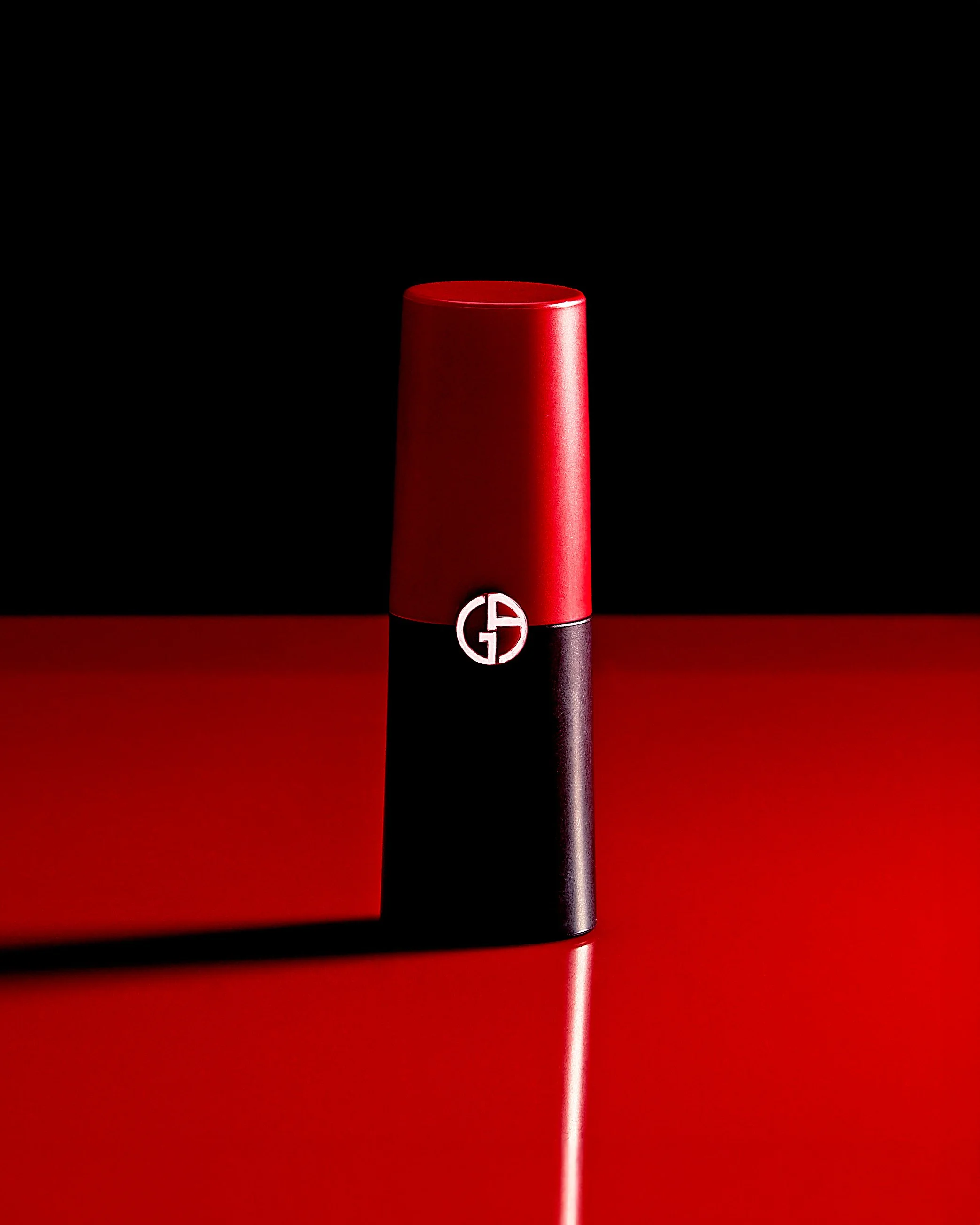 A red lipstick with a black and silver base, standing upright on a red surface against a dark background.