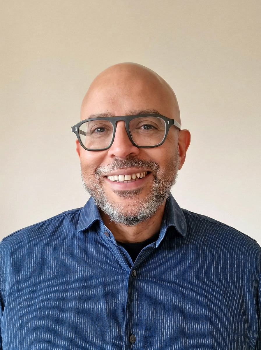A smiling middle-aged man with a bald head, gray beard, glasses, wearing a blue striped button-up shirt against a plain beige background.