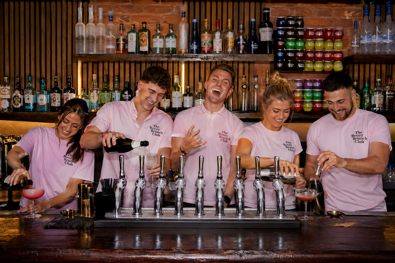 Bar staff in pink shirts pouring and preparing cocktails behind a bar with liquor bottles and colorful jars in the background.