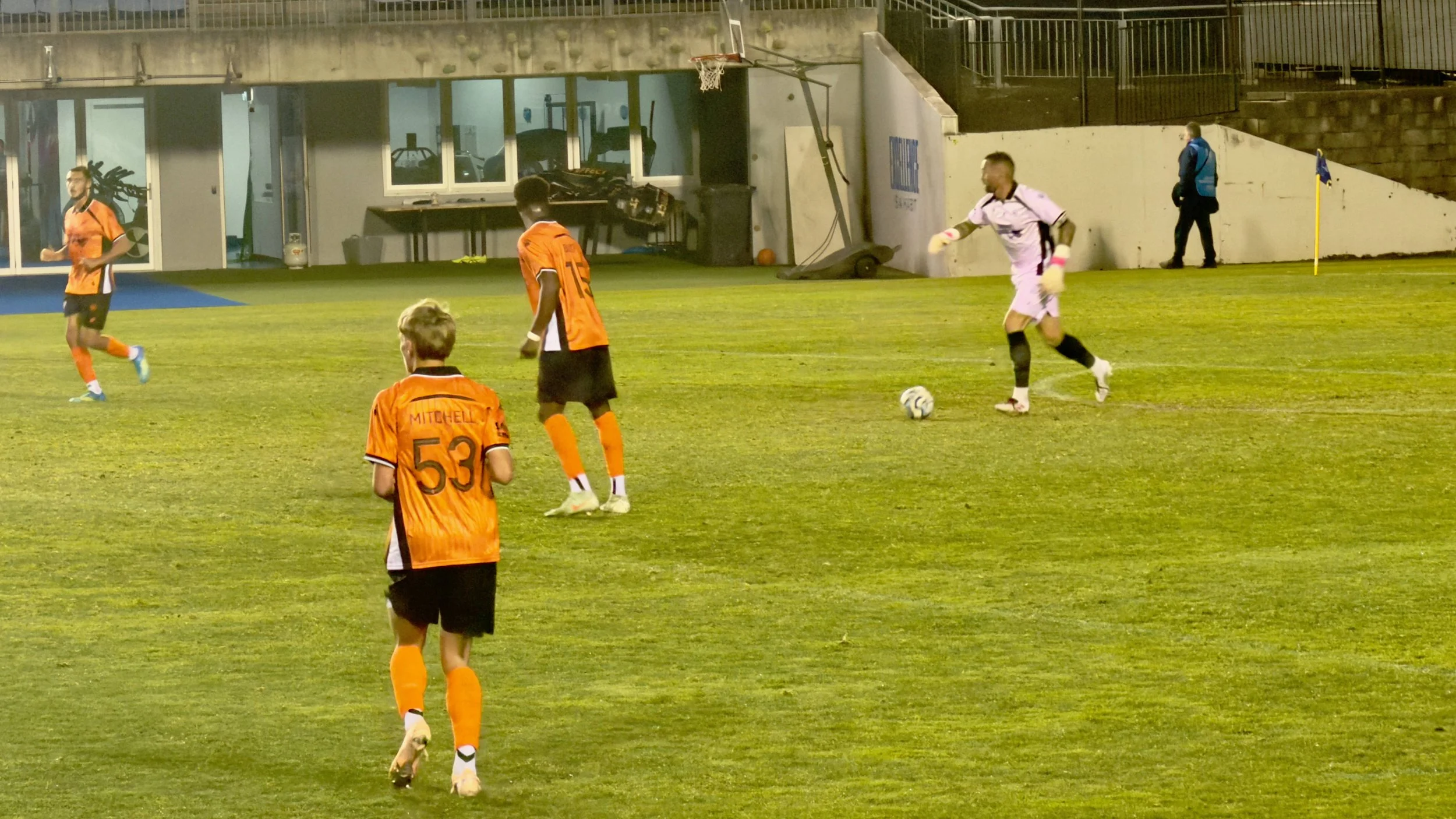 Brisbane Roar players in orange spread across the pitch as a teammate controls possession, with a Sydney Olympic opponent approaching at Belmore Sports Ground.
