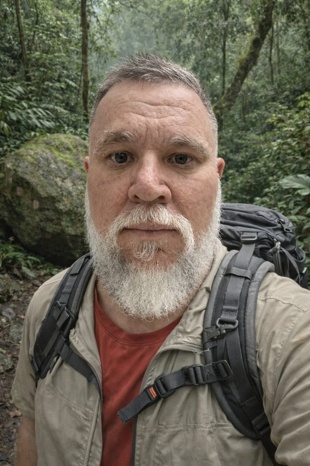 A man with a beard and short hair taking a selfie in a dense green forest, wearing a beige jacket and a backpack.
