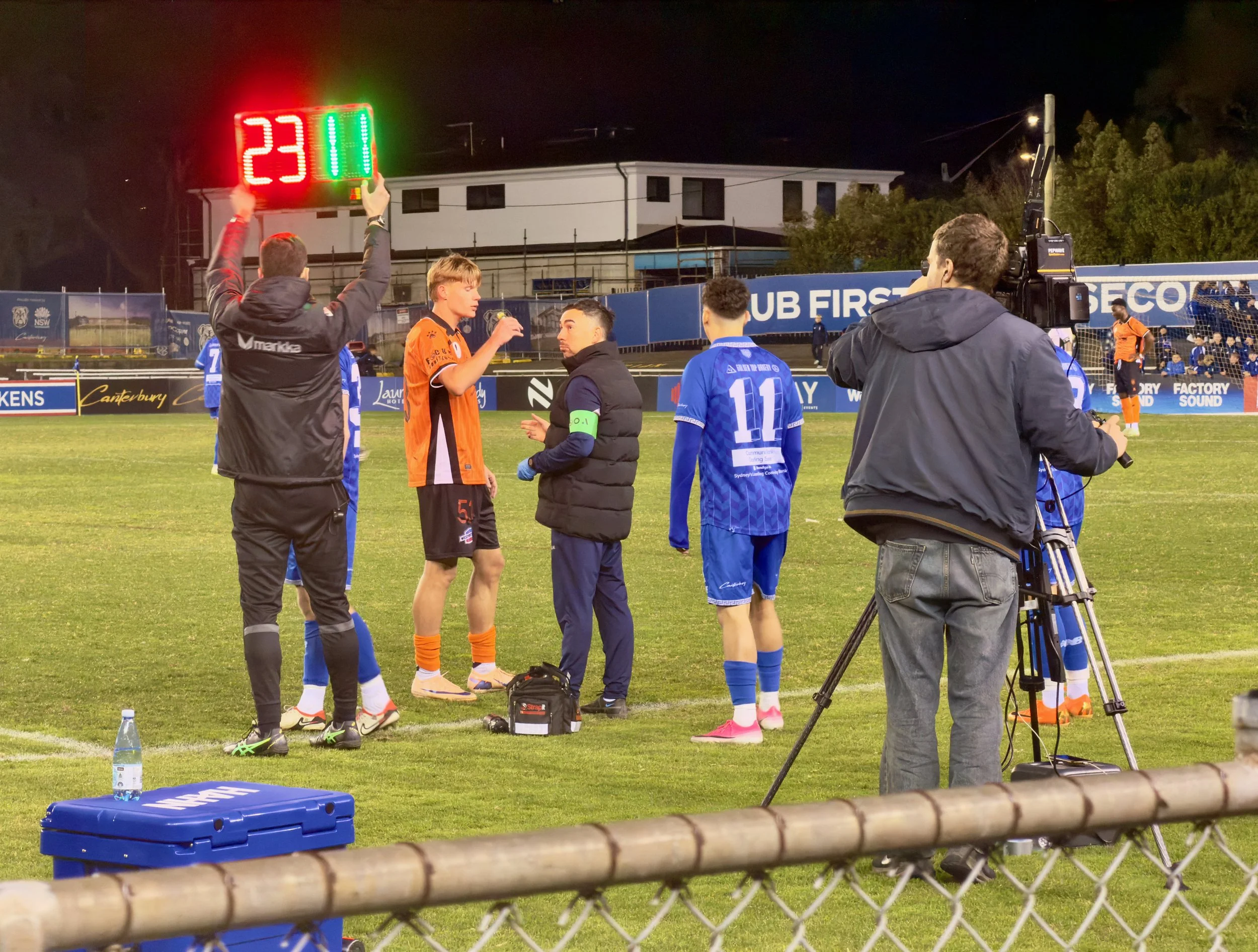 A fourth official holds an electronic substitution board showing 23 and 11 while players and broadcast staff wait on the Belmore Sports Ground touchline.