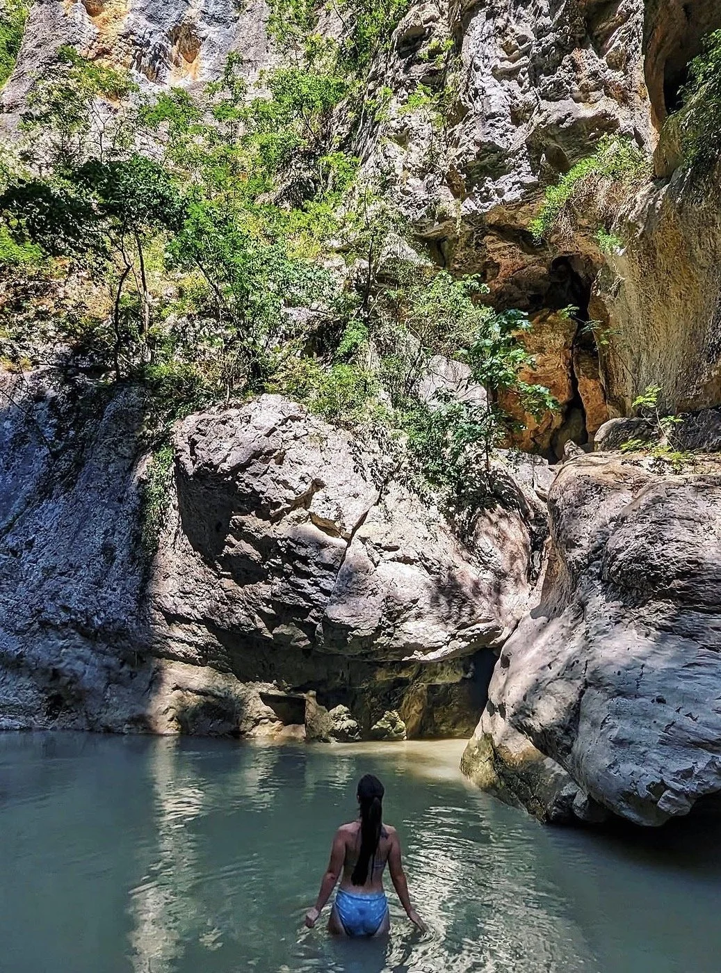 A woman with long dark hair, wearing a blue swimsuit, wading in a natural pool surrounded by large rocks and greenery, with a towering canyon wall in the background.