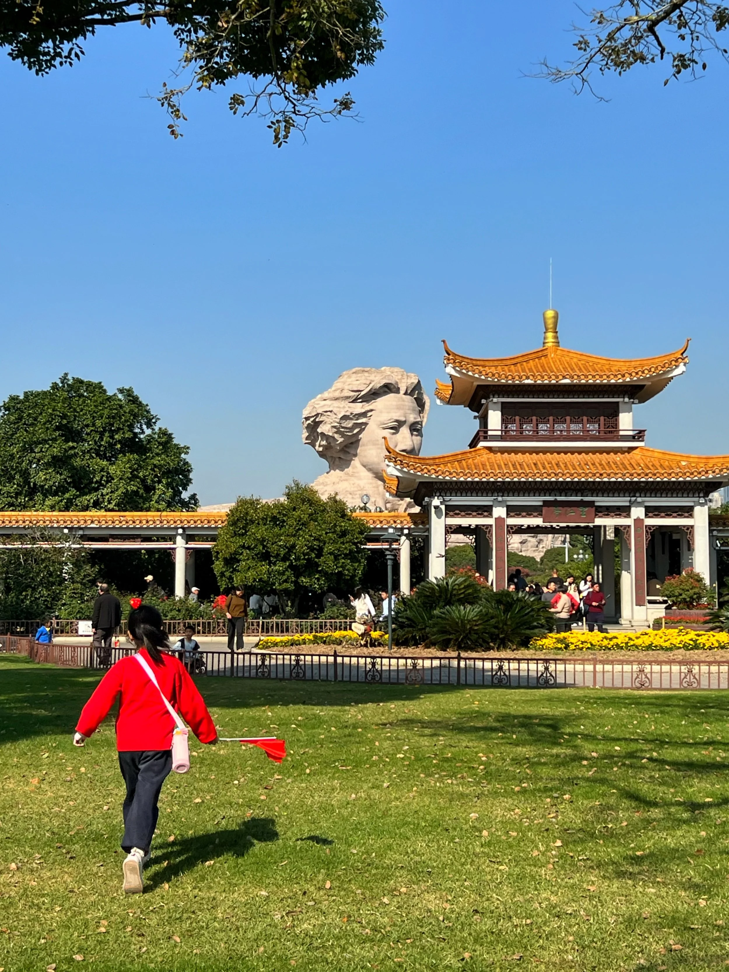 A park scene with a woman in a red jacket walking on green grass carrying a pink mat and red flag, with trees, a traditional Chinese-style pavilion, and a large stone sculpture of a woman's face in the background under a blue sky.