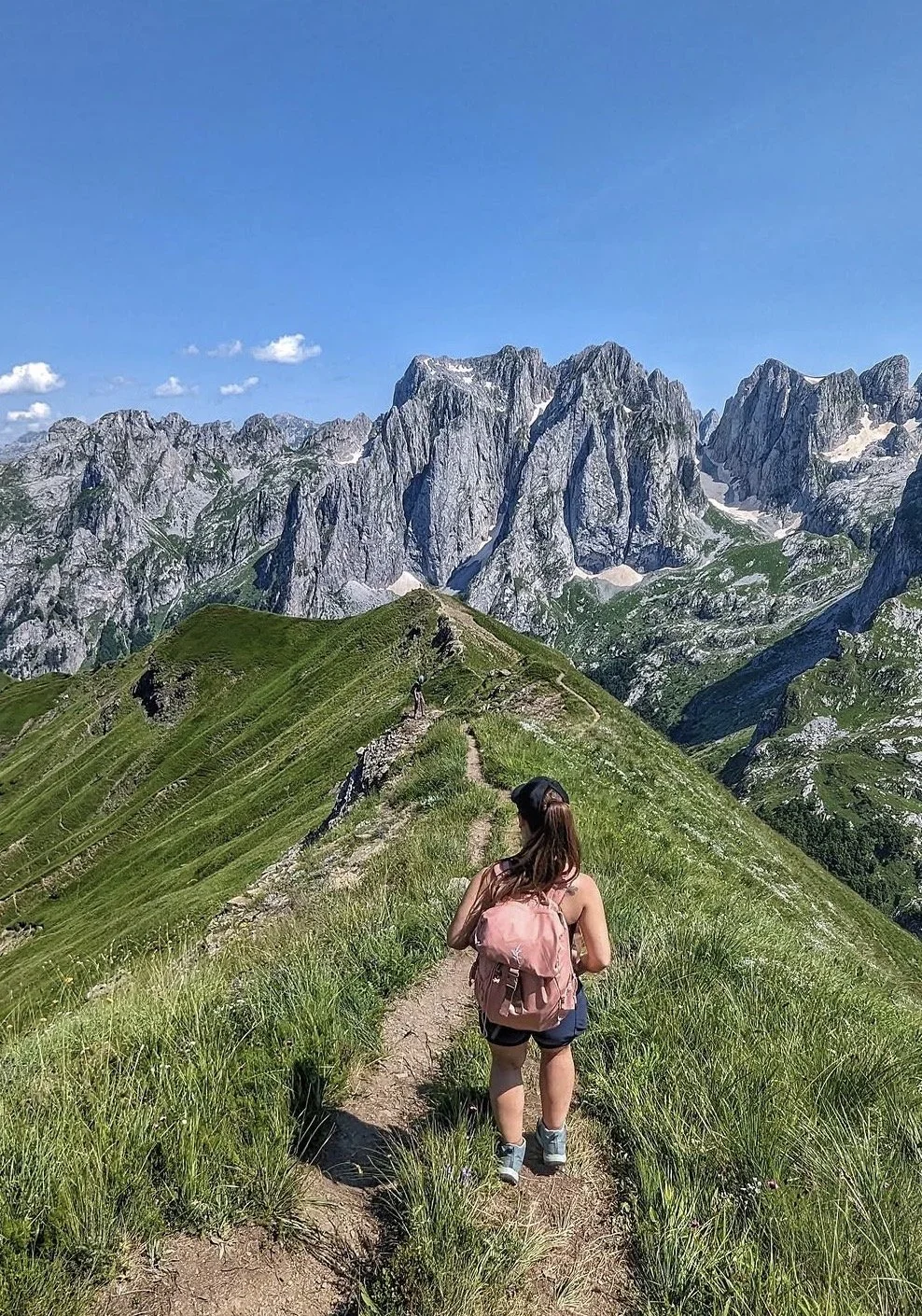 A woman hiking on a narrow dirt trail along a grassy mountain ridge with tall rocky mountains in the background under a clear blue sky.