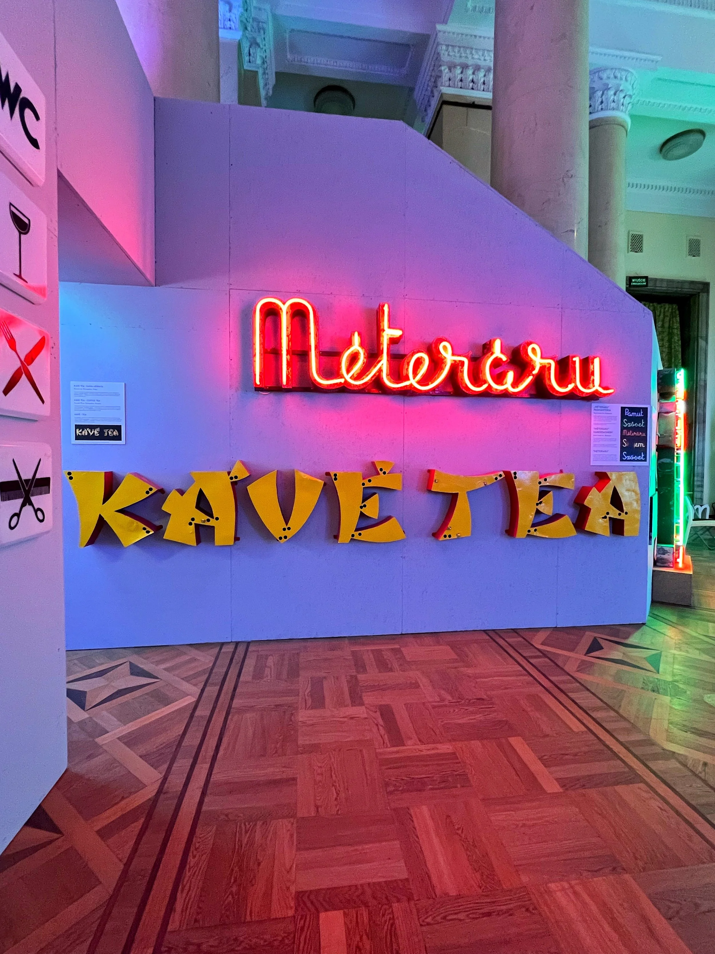 A brightly lit neon sign spelling 'Metoru' hangs on a wall with pink and red glow. Below it, large yellow letters spell out 'KAVE TEA' against a light-colored wall.