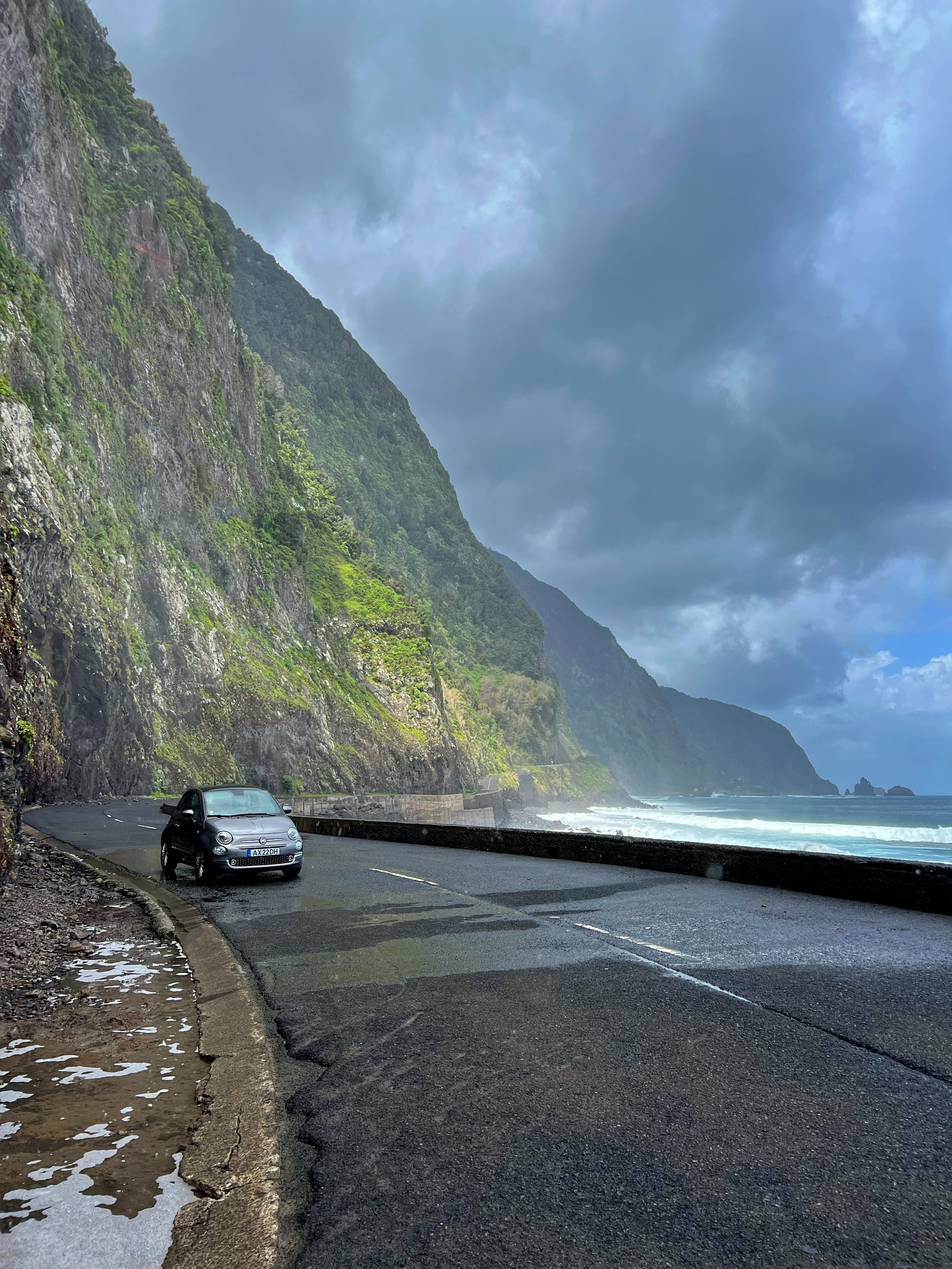 A scenic coastal road running alongside the ocean, with a black car parked on the side. The road is wet, with drifted snow along the edge. Tall, lush green cliffs rise steeply on the left, with a cloudy sky overhead.