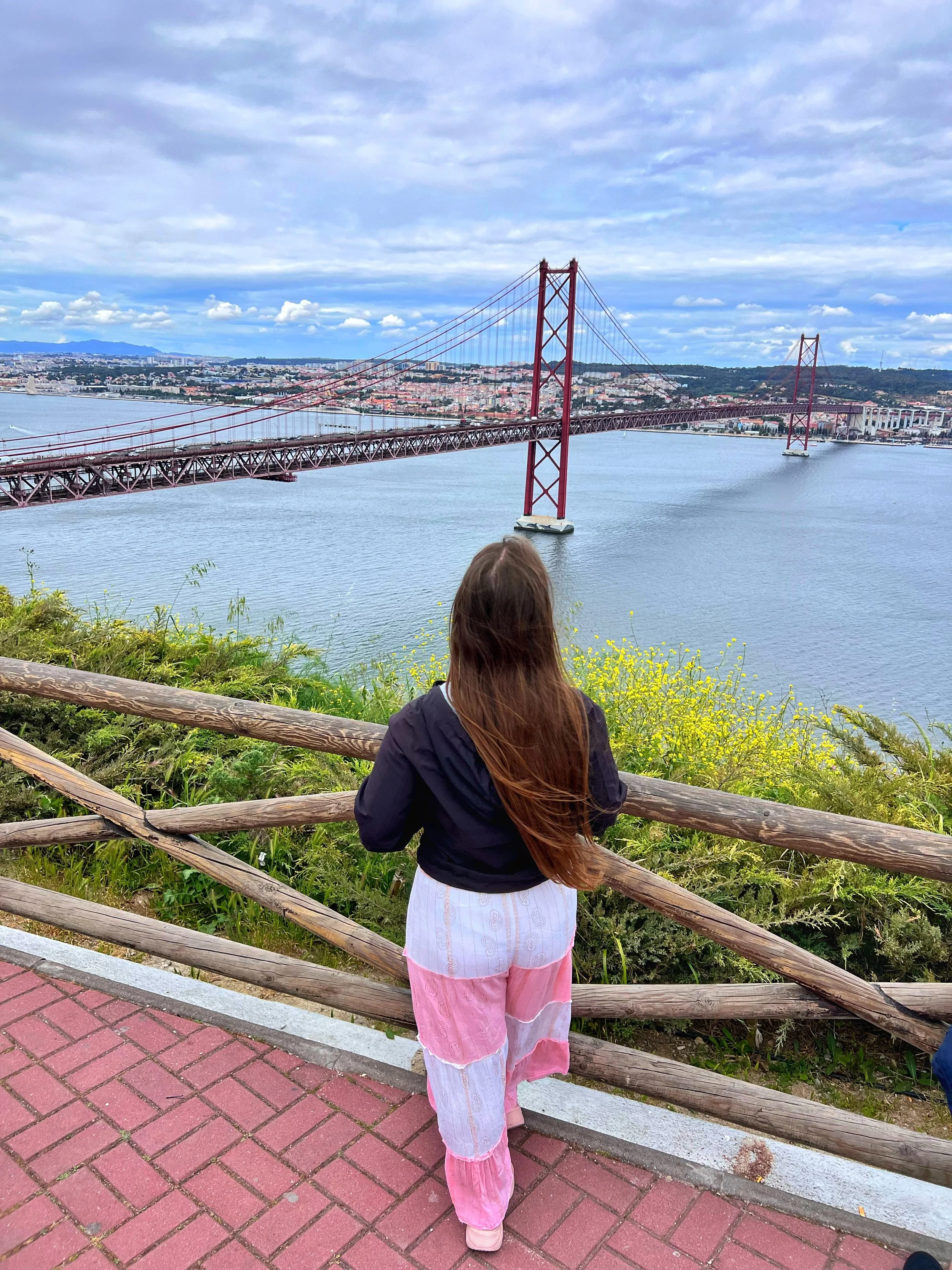 A person with long brown hair wearing a dark jacket and pink and white pants stands facing away, looking at a large bridge over a body of water with a cityscape in the background.