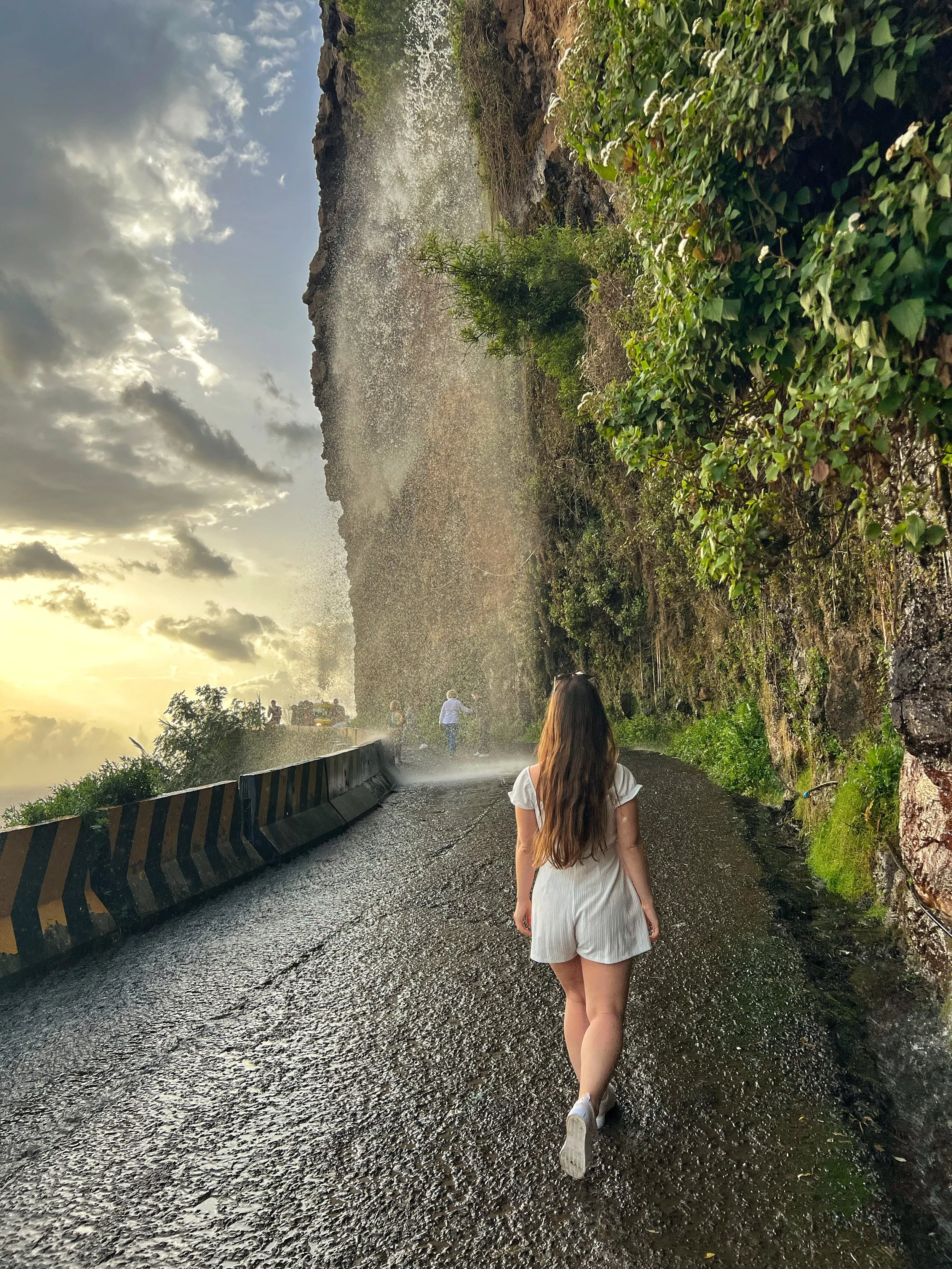A woman with long brown hair wearing a white dress, walking along a wet, rocky road with a guardrail on the left. There are other people in the distance walking near a mist or spray, with a cloudy sky and some sunlight breaking through.