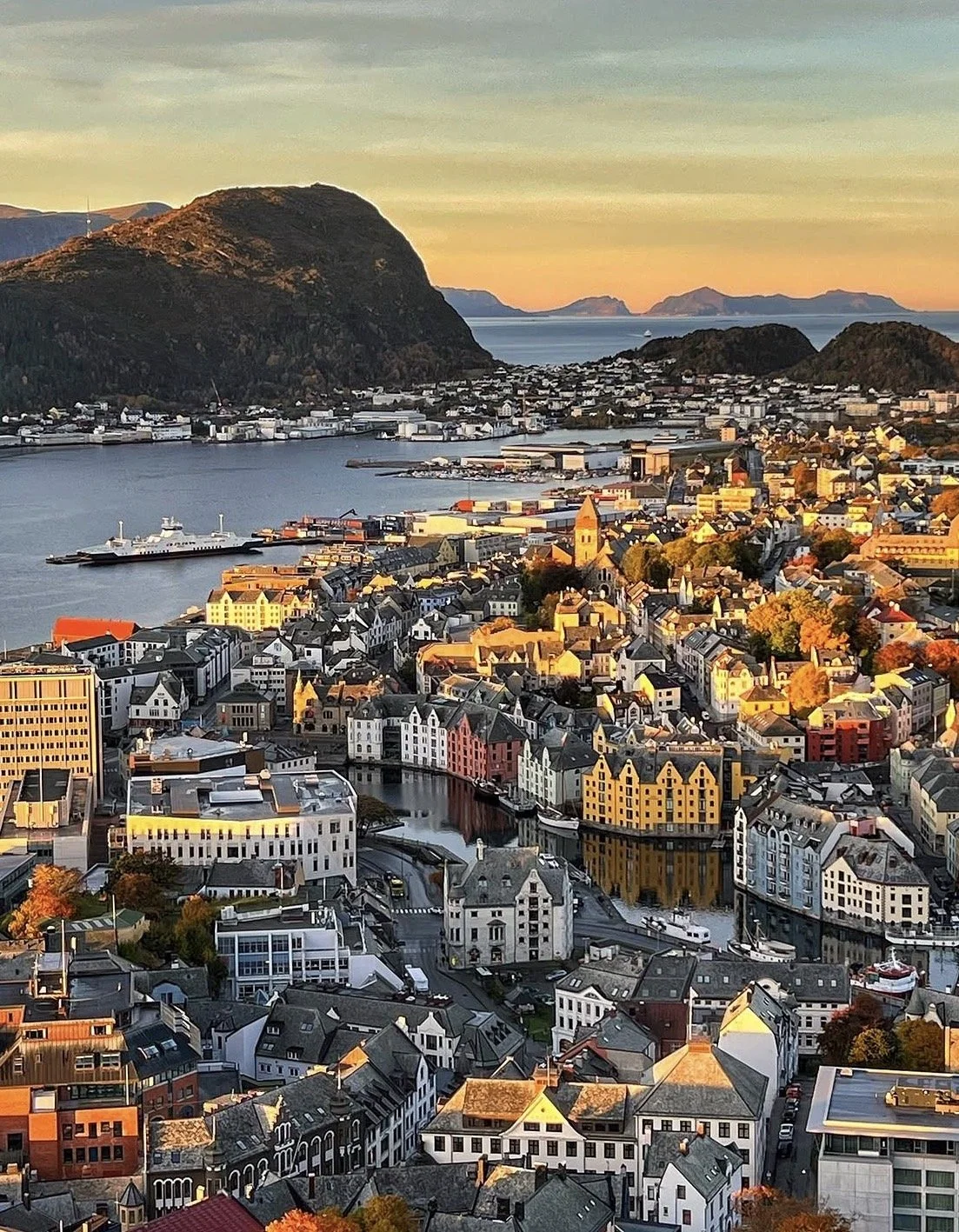 Aerial view of a coastal city with colorful buildings, a harbor with ships, and mountains in the background during sunset.