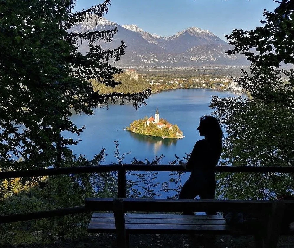 A woman stands on a viewing platform overlooking a lake with an island church, surrounded by trees, mountains, and a clear sky.