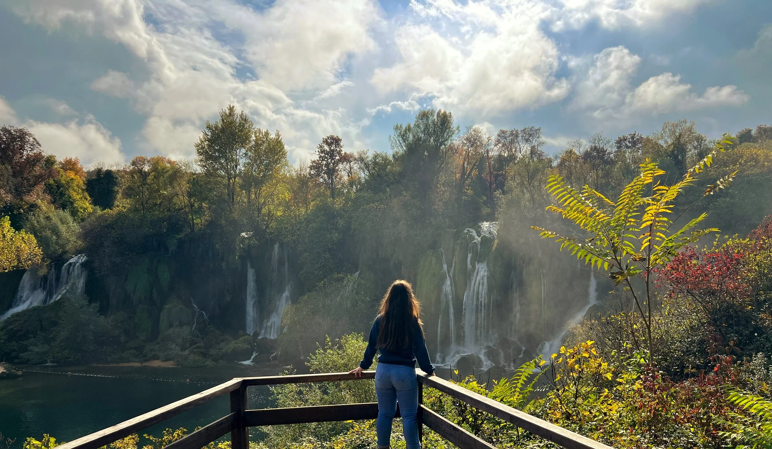 A woman with long hair stands on a wooden viewing platform overlooking a scenic waterfall surrounded by dense autumn trees, with clouds and a blue sky above.