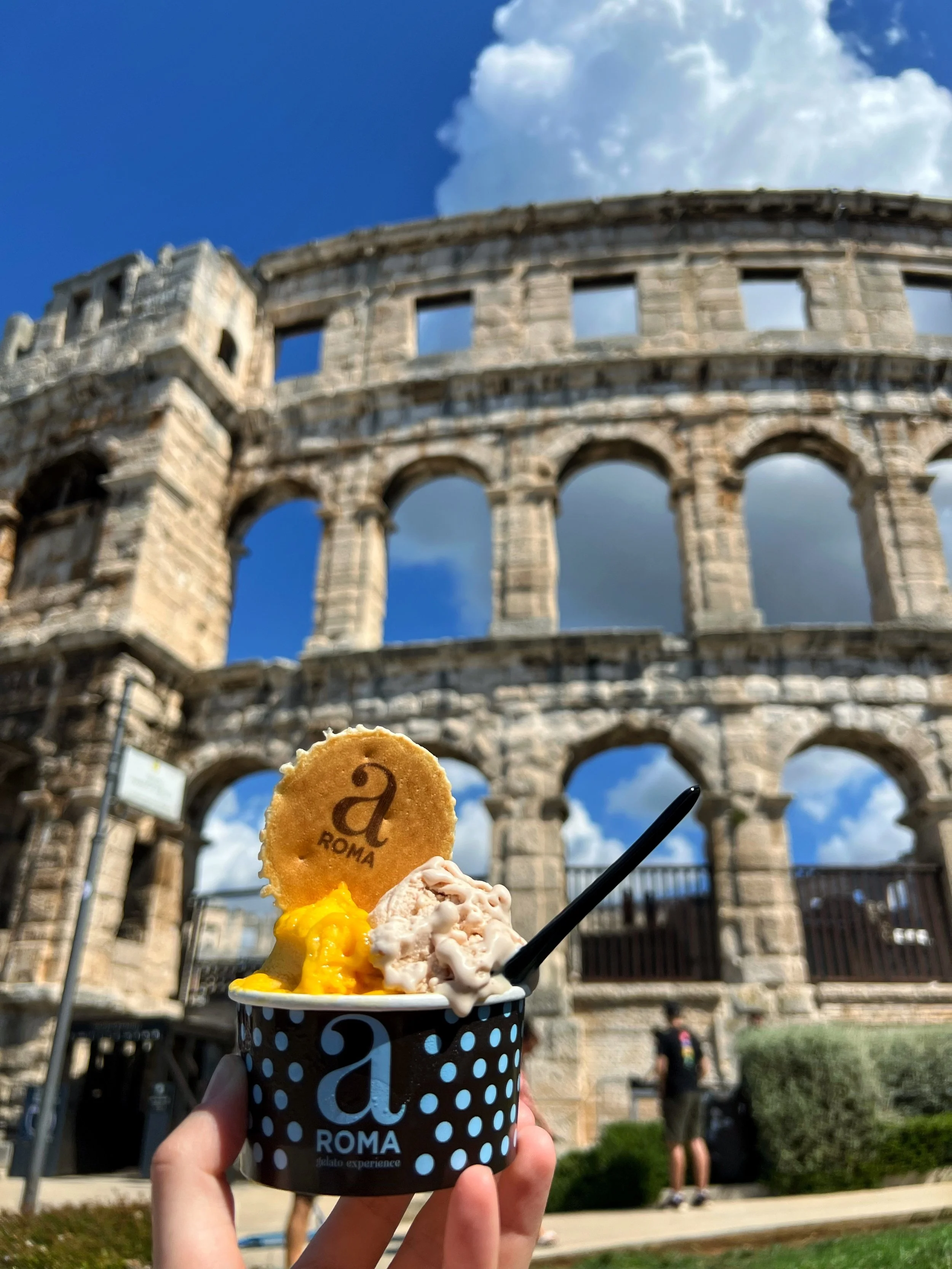 A cup of gelato with a cookie that says "ROMA" and a black spoon, held in front of the Roman Colosseum against a blue sky with some white clouds.