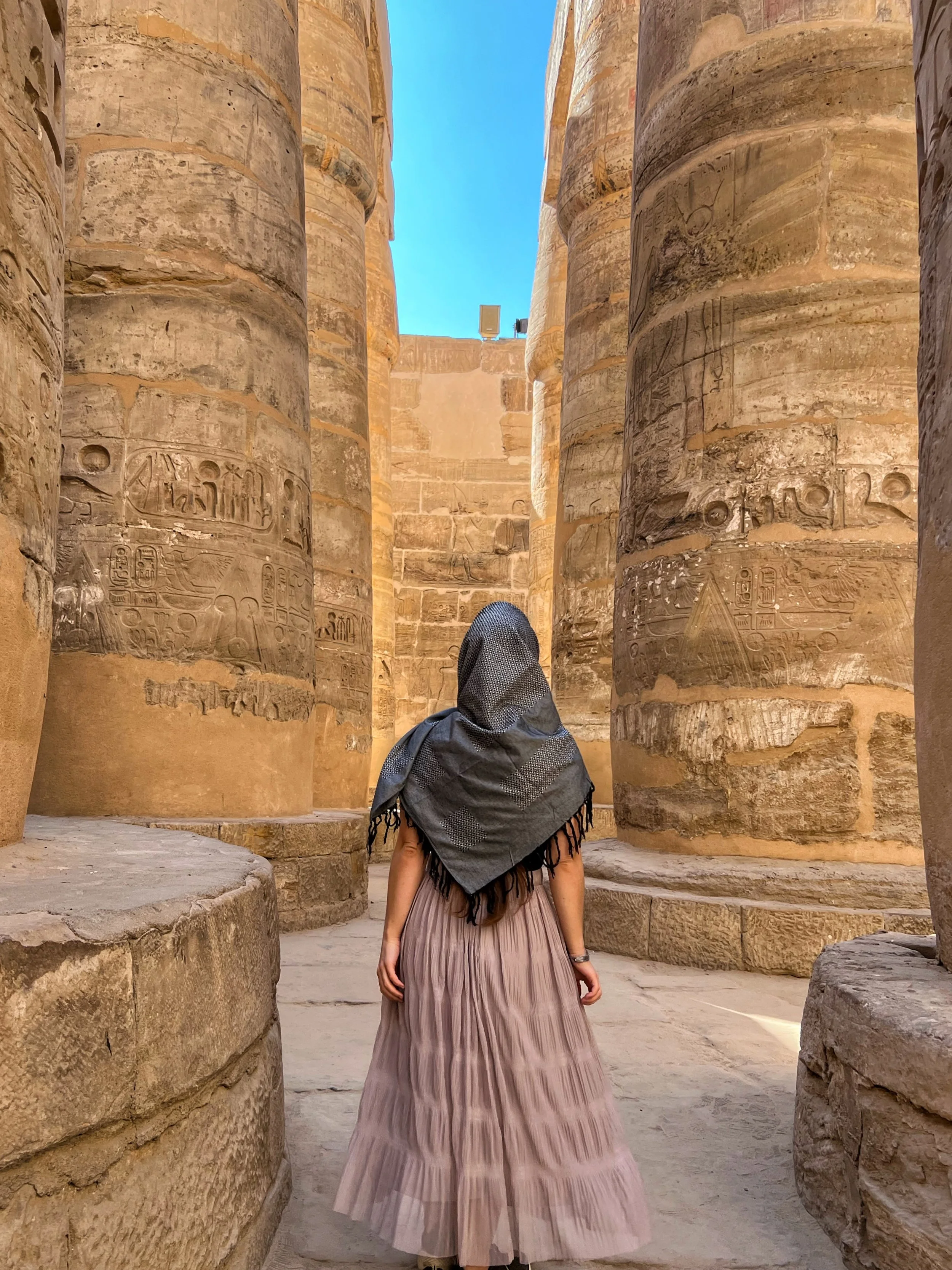 A woman standing between massive ancient stone columns with hieroglyphs carved into them, looking towards a brightly lit, open sky.