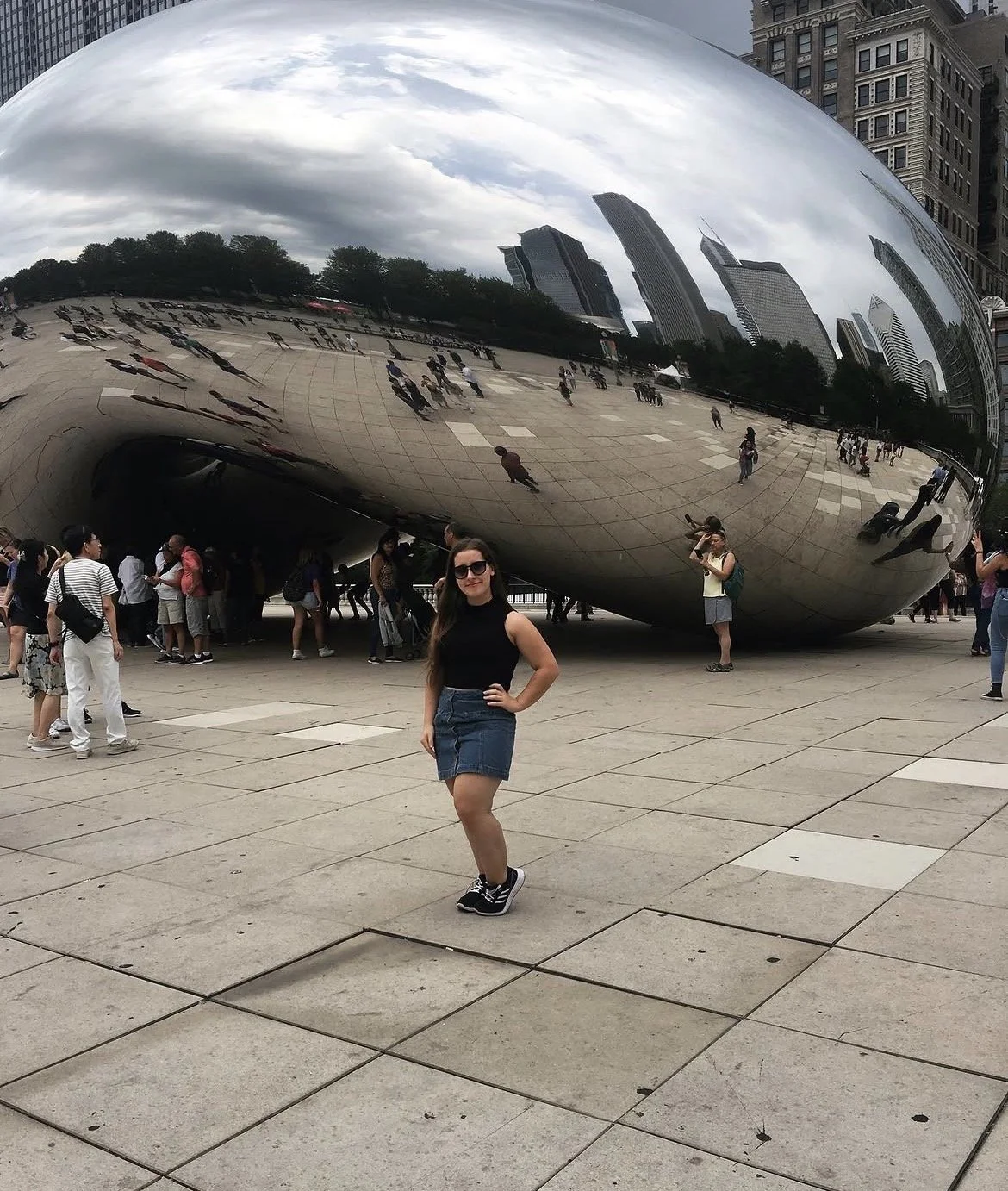 A woman with sunglasses, black sleeveless top, denim skirt, and sneakers stands in front of the Cloud Gate sculpture in Millennium Park, Chicago. The reflective, bean-shaped sculpture mirrors the city skyline and cloudy sky, with other visitors around.