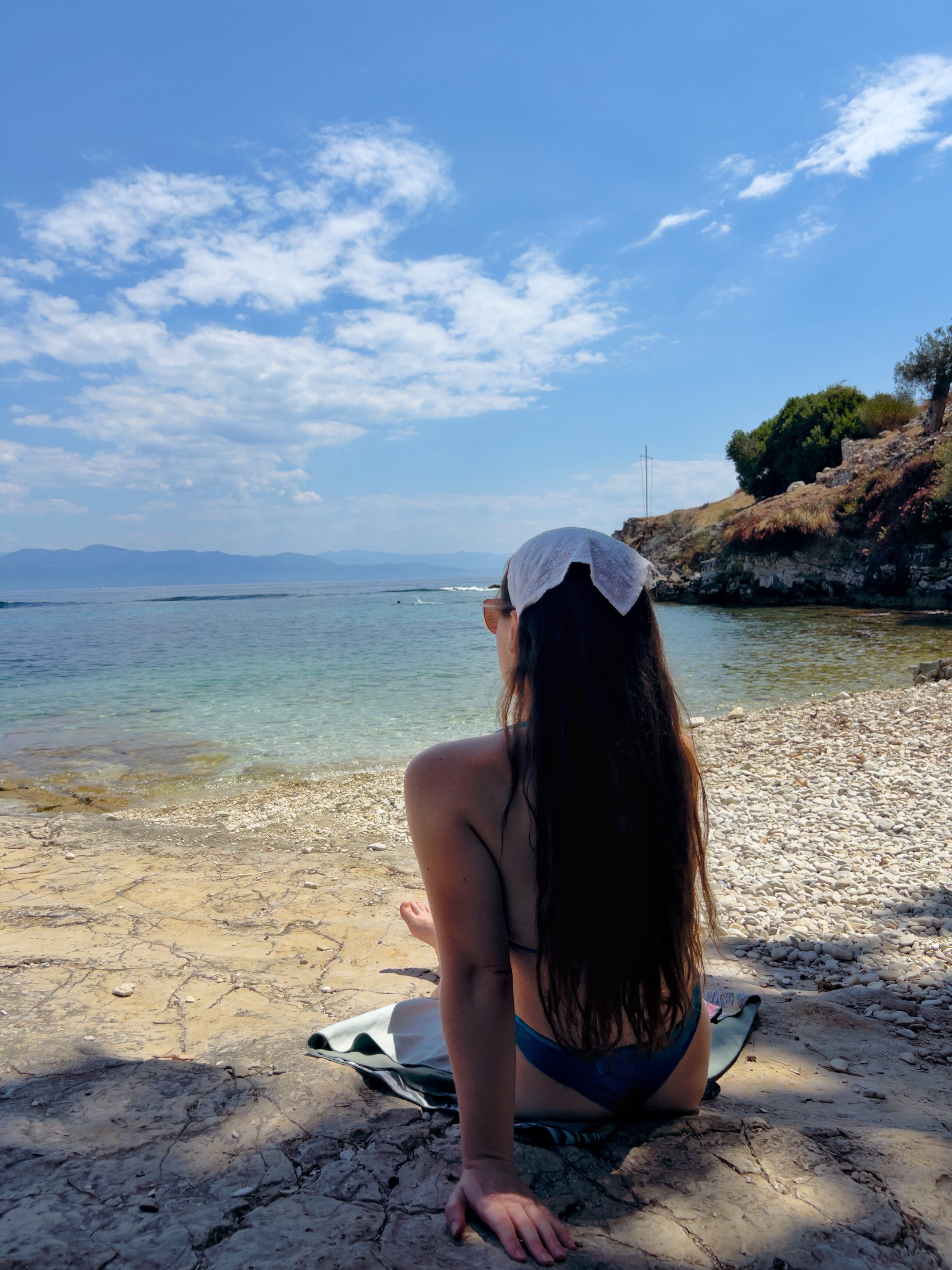 A woman with long dark hair sitting on a rocky beach, facing the water with a view of the ocean, hills, and sky with scattered clouds.