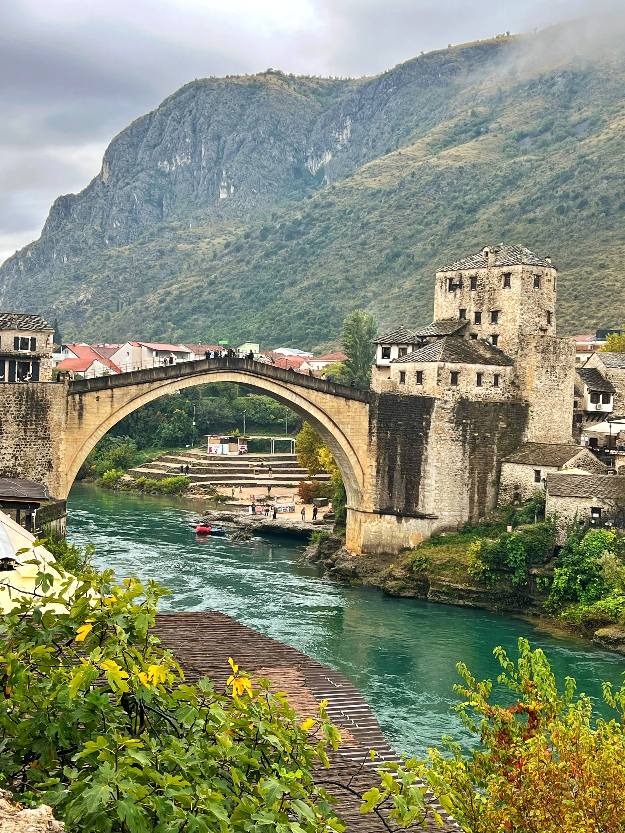 A scenic view of a historic stone bridge and castle-like building over a river, with lush greenery and mountains in the background, and cloudy sky overhead.