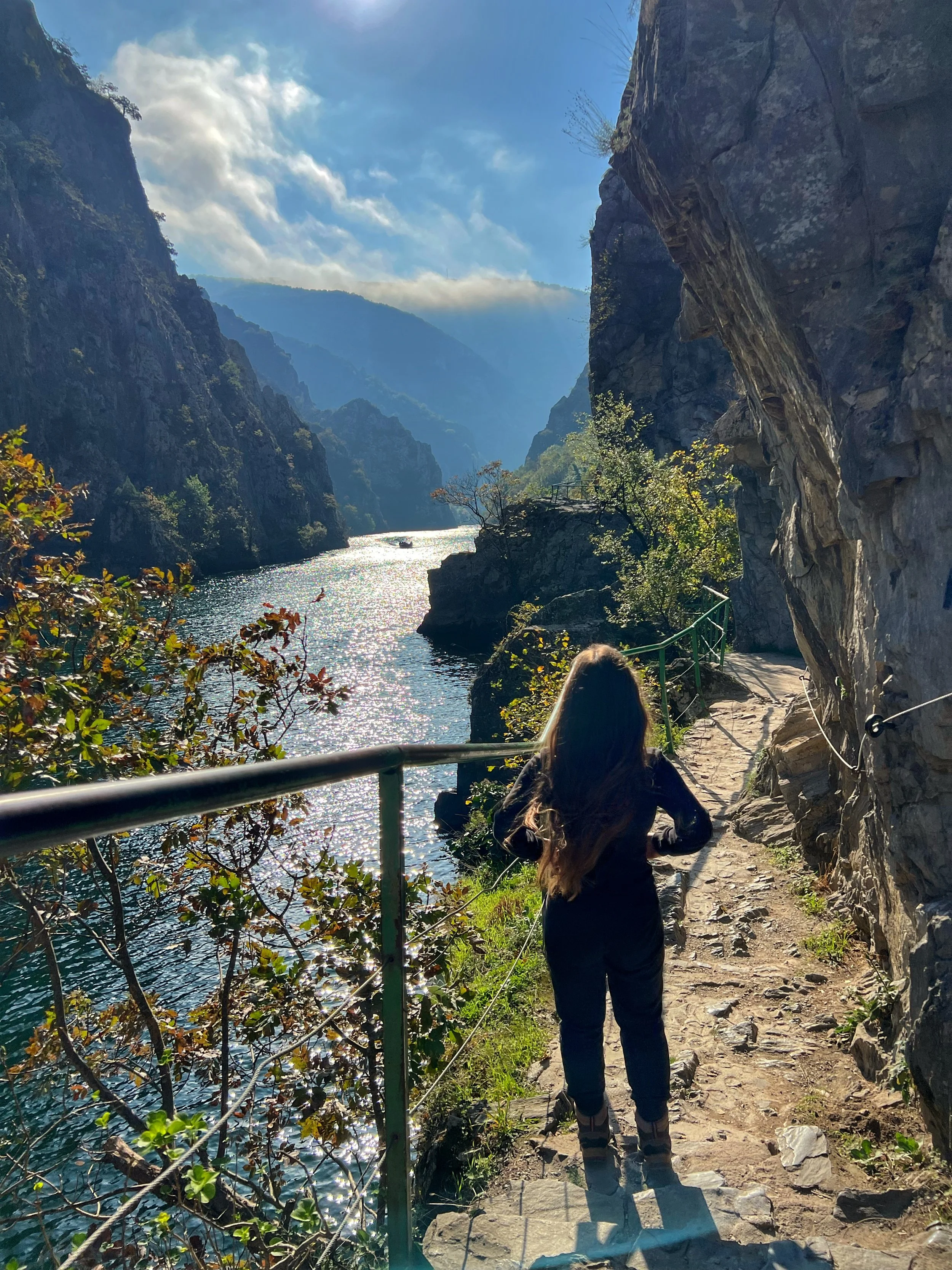A person standing on a trail next to a rocky cliff, overlooking a river surrounded by mountains under a blue sky with clouds.