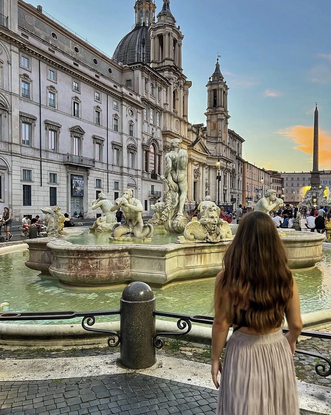A woman with long brown hair wearing a beige skirt stands in front of the Fountain of the Four Rivers in Piazza Navona, Rome, Italy, during sunset, with historic buildings and the obelisk in the background.