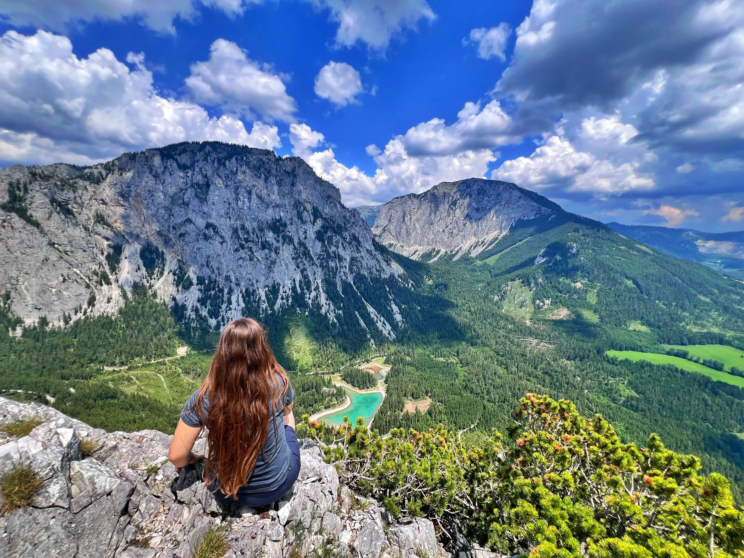 A person with long brown hair sitting and looking out over a scenic mountain landscape with green valleys, rocky ridges, a small turquoise lake, and a partly cloudy sky.